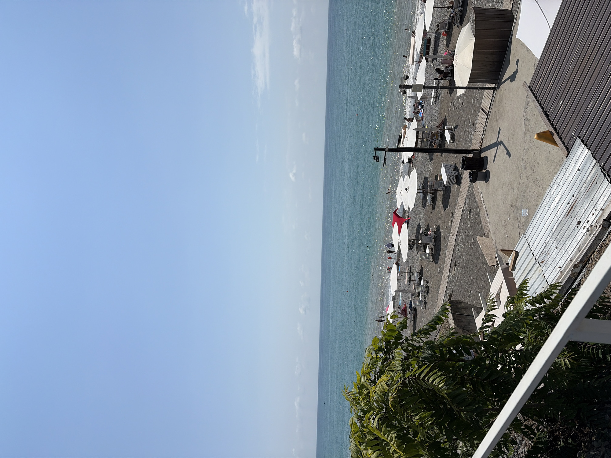 The image captures a view of a pebble beach in Sochi, Russia, on a sunny day. The sea is a calm, turquoise blue, meeting a wide expanse of sky that is mostly clear with a few wispy clouds. The beach is dotted with numerous white and red umbrellas, providing shade for beachgoers. People are visible lounging on beach chairs, walking along the shore, and wading in the shallow water. Wooden pathways run across the beach, connecting different areas. In the foreground, lush green foliage from a tree hangs into the frame, and a white railing is visible. Structures such as beach huts, trash cans, and lamp posts are also present on the beach. The overall scene depicts a typical day at a seaside resort.