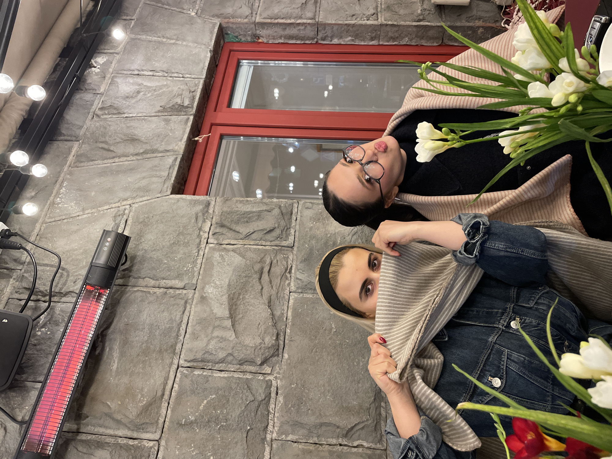 Two women are posing for a photo outdoors. One woman with dark hair and glasses is facing the camera and pouting, while the other, wearing a black headband and a denim jacket, is partially covering her face with a striped scarf. Both are wearing jackets, and white flowers are next to them. They appear to be sitting next to a stone wall with a red-framed window and an outdoor heater. Based on the outdoor heater, the location is in Yerevan, Armenia.