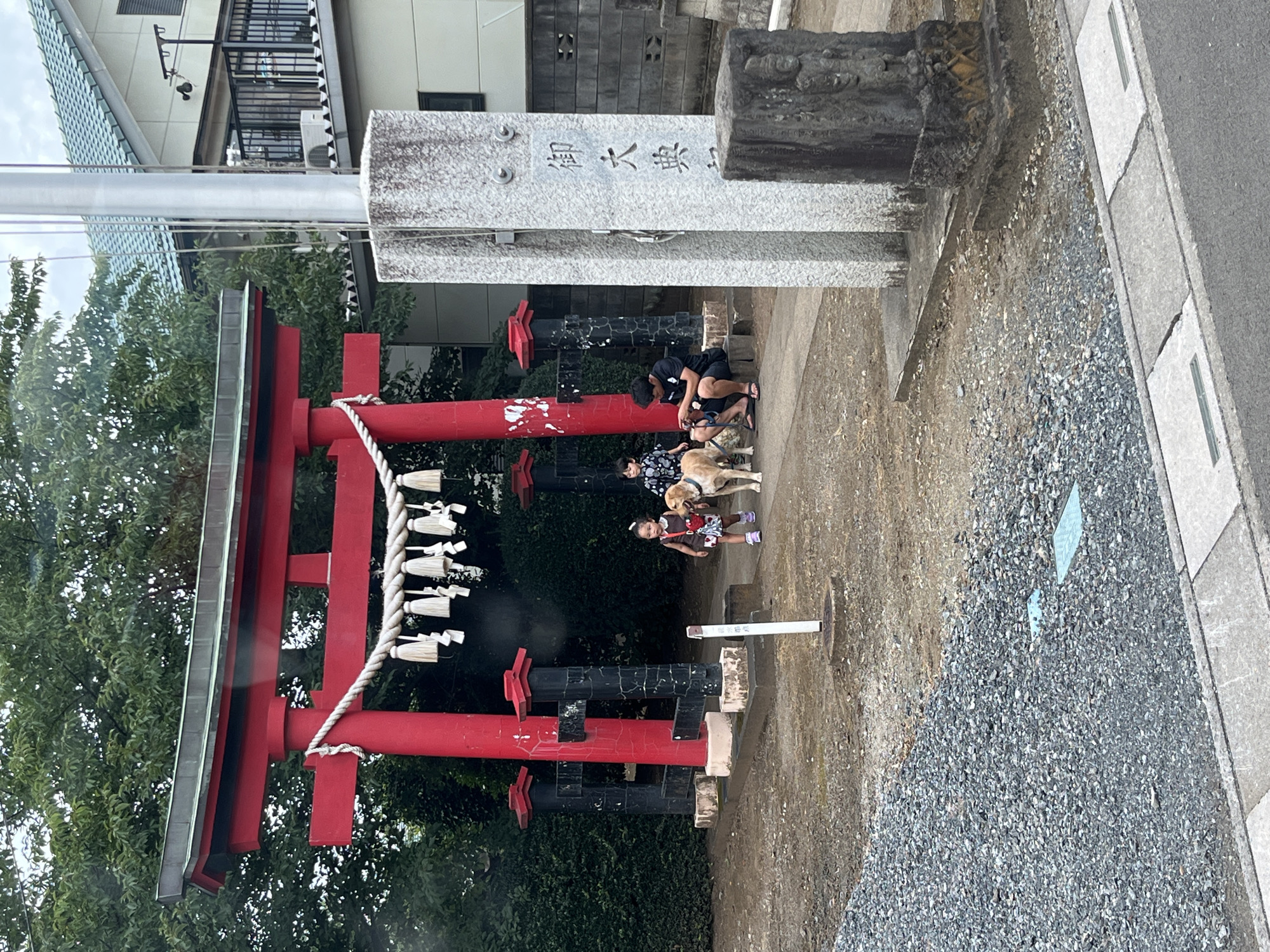 This image depicts a scene at a shrine in Sayama, Japan. In the foreground, a person is seated on the ground next to a light-colored dog. To their left, a young child walks with another dog. Behind them, a large red Torii gate stands, adorned with white tassels. The shrine appears to be located in a somewhat urban or suburban area, with houses visible in the background. The ground is covered with gravel and dirt. There are stone monuments present, one of which has Japanese calligraphy. The weather appears overcast.
