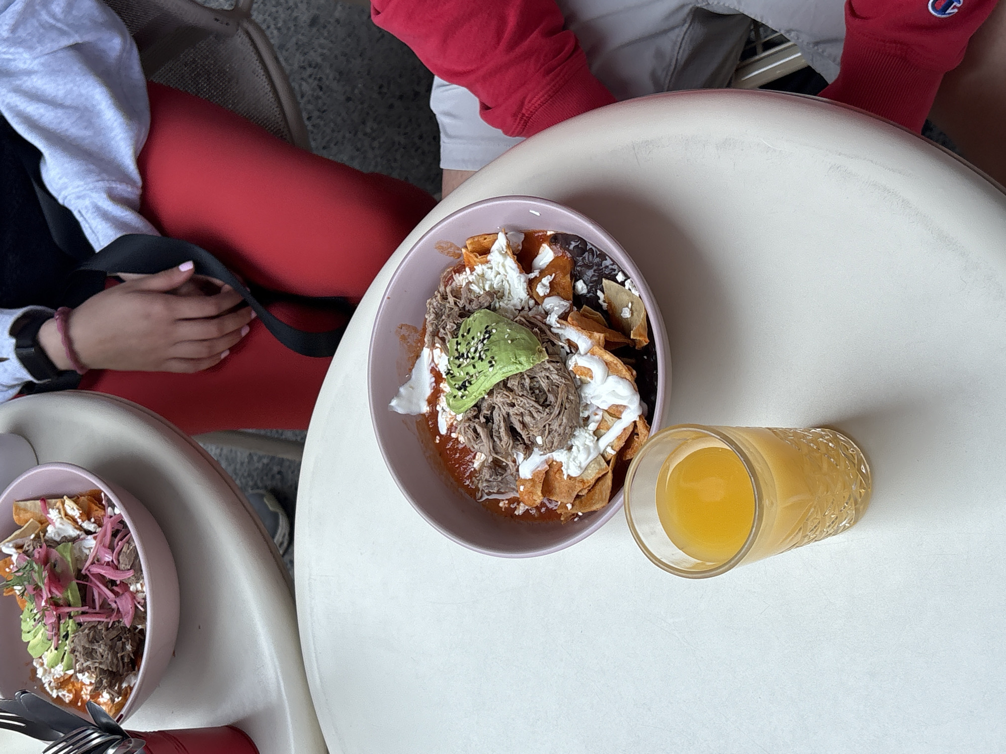 This is an overhead shot depicting a casual dining scene, likely at an outdoor cafe or restaurant in Puebla, Mexico. The image focuses on two tables with food and drinks, with two individuals partially visible.
In the foreground, a light-colored, round table holds a prominent pink bowl filled with chilaquiles. This dish consists of tortilla chips generously coated in a red sauce, topped with shredded meat (appearing to be pork or beef), black beans, crumbled white cheese (queso fresco or cotija), a drizzle of sour cream or crema, and sliced avocado garnished with black sesame seeds. Beside this bowl, a textured clear glass holds a yellow-orange beverage, likely orange juice.
To the bottom left, another similar light-colored table features a second pink bowl of chilaquiles. This portion also includes shredded meat and avocado, along with what appears to be pickled red onions and fresh herbs. Silverware is visible near this bowl.
One person is seated on the left, wearing a light grey long-sleeved sweatshirt and red pants or leggings. Their left hand, with light pink nail polish, rests on a black strap, possibly from a bag or a chair. A dark-colored watch or bracelet is on their wrist. Another person is partially visible on the upper right, wearing a red long-sleeved shirt with a white, circular logo on the cuff, possibly a Champion logo, but it is not fully discernible.
The overall scene suggests a pleasant daytime meal, possibly breakfast or brunch, given the type of food and drink. The tables are light-colored, and the background shows hints of outdoor seating with grey textures, possibly concrete or tiled flooring and metal chair frames.