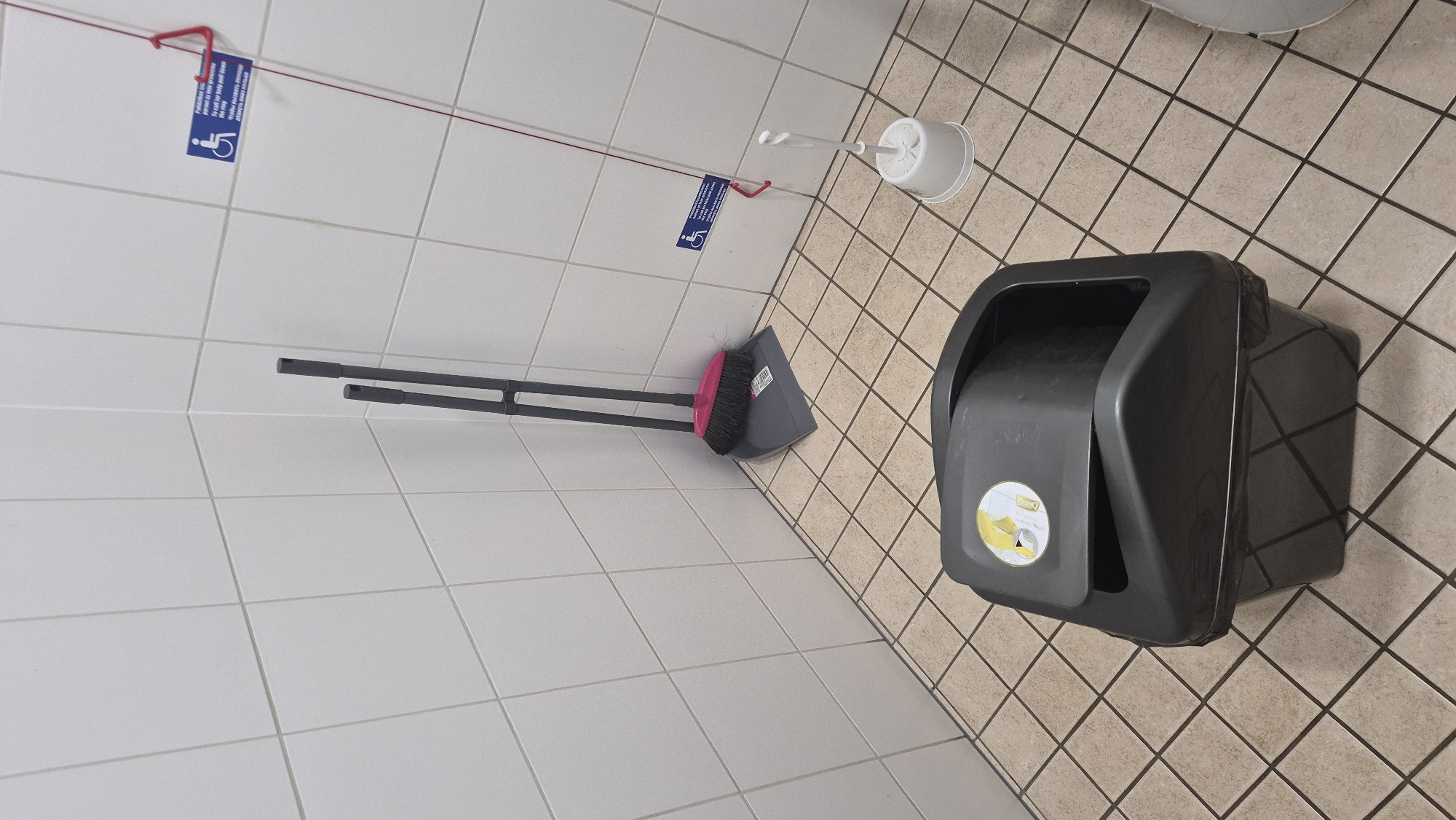 The image depicts a corner of a room, likely a public restroom in Dagda, Latvia, characterized by tiled walls and floor. In the foreground, a large, dark gray trash can with a hinged lid sits prominently. The lid is partially open, revealing a black trash bag lining the interior. A circular graphic is visible on the lid, featuring yellow and white elements.
To the left of the trash can, leaning against the wall, is a dustpan and brush set. The dustpan is gray, and the brush has pink bristles and a gray handle. Behind the dustpan, a long, thin red cord extends diagonally upwards towards the top right of the frame.
On the wall in the upper right quadrant, a blue and white sign with an accessibility symbol is affixed. Red plastic clips are attached to the top of this sign, and the red cord appears to be threaded through these clips. Further to the right, a white toilet brush sits in its holder on the floor. The overall impression is a utilitarian space, possibly a public restroom in Dagda, Latvia, containing cleaning supplies and waste disposal.