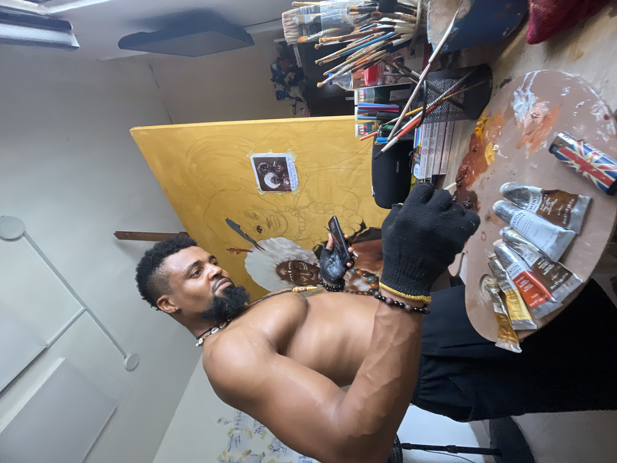 A shirtless Black man stands in what appears to be an art studio. The setting has the appearance of an artistic haven, filled with numerous brushes and various art supplies, including paint tubes, a palette covered in mixed paints, and several books. In the background, there's a large yellow canvas featuring an incomplete portrait. The man is holding a phone, wearing a black glove on one hand and beaded bracelets on both wrists. He wears a beard and a beaded necklace. It is not possible to know if the location is Ado, Nigeria.