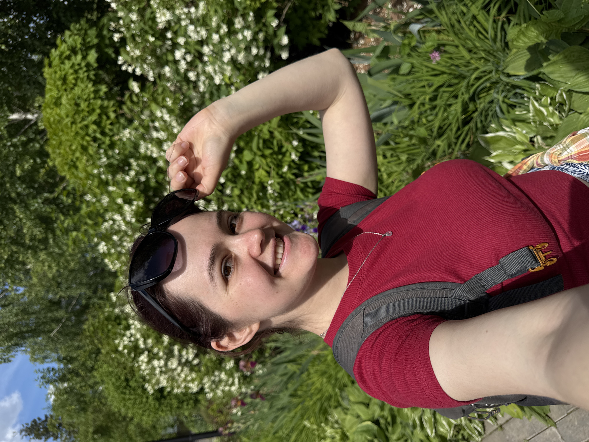 A young adult female is depicted in a close-up, chest-up selfie in an outdoor, verdant environment. She possesses fair skin with visible freckles across her cheeks and nose, dark eyes, and dark brown hair pulled back from her face, with sunglasses resting on her head. Her right hand is raised, lightly gripping the frame of the black, slightly cat-eye style sunglasses. She wears a form-fitting, short-sleeved, ribbed red top and a silver necklace with a single clear or white teardrop-shaped pendant. The grey straps of a backpack are visible over her shoulders, with an orange/gold buckle on the right strap. A patterned, multi-colored fabric is visible on her lower right side. She is smiling directly at the camera, with teeth visible.
The background consists of dense green foliage, indicating a park or garden setting. Lush green trees and shrubs are prominent, with numerous small white flowers blooming on several bushes, suggesting a species like Spiraea. Various other green leafy plants, including hostas, and hints of purple or pink flowers are visible at mid-ground. A patch of blue sky with white clouds is seen in the upper left corner, while a paved pathway is partially visible on the lower left. The lighting is bright and sunny, casting a clear illumination on the subject and environment. The image provides no specific visual cues or landmarks to definitively identify the location within the city of Izhevsk, Russia.