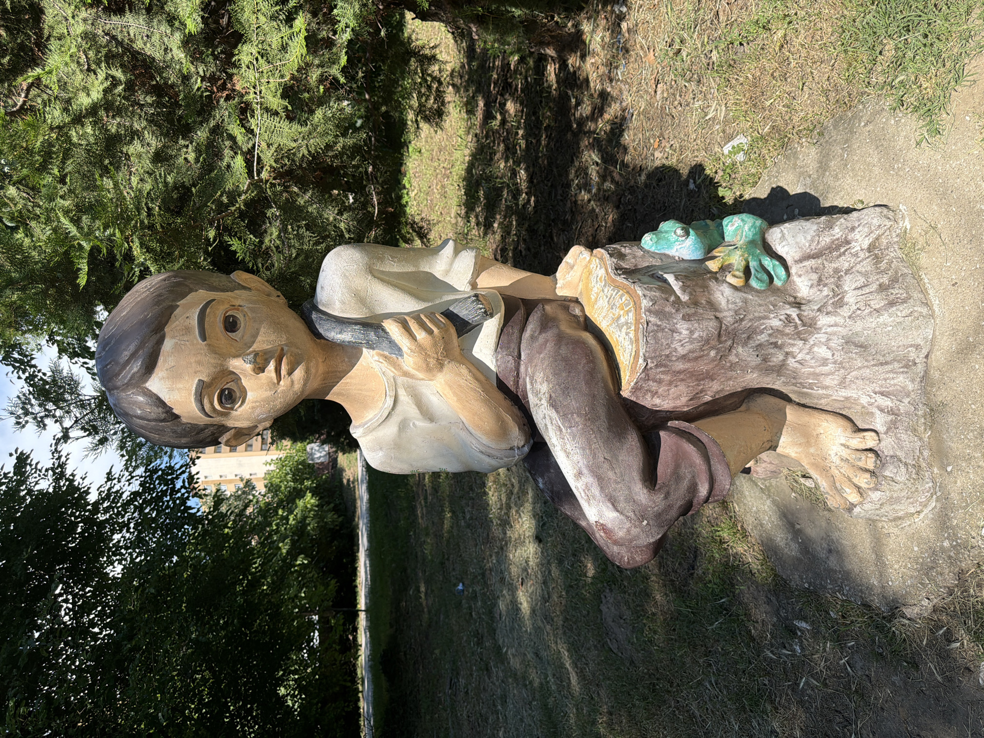 A statue of a young boy sitting on a tree stump is positioned in an outdoor park setting. The boy has dark hair, large eyes, and is wearing a light-colored shirt and brown pants. He is holding a dark object, possibly a stick or tool, in his right hand. A small, green frog statue is attached to the side of the tree stump. The ground is covered with grass and dirt, and trees are visible in the background, along with a building. The image appears to be taken in Pazardzhik, Bulgaria.
