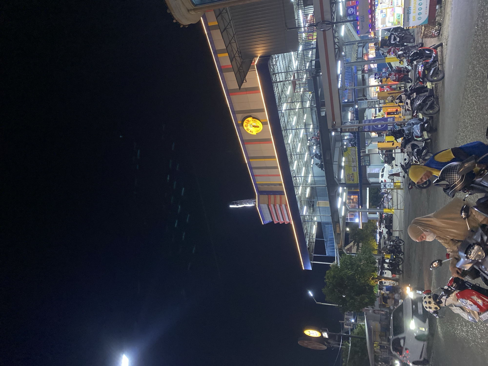 At night in Lhokseumawe, Indonesia, a brightly lit commercial structure houses a business with a circular yellow logo reading "MR. D.I.Y.". Parked motorcycles fill the foreground and side area in front of the building. One figure, possibly a female, is seen wearing a hijab and mask, sitting on a motorbike. Street lights illuminate the area, casting light across the road and towards the sky. The Indonesian flag is displayed on the building.