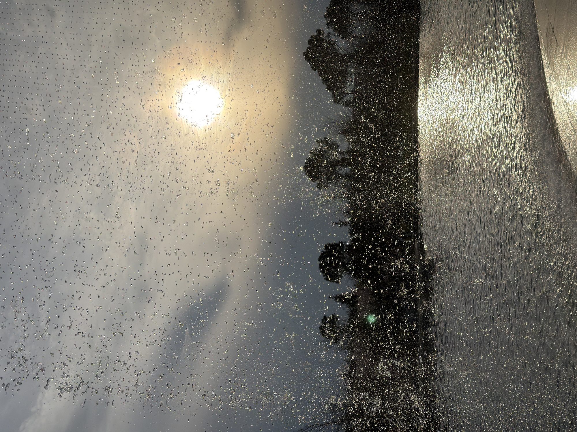 The image captures a perspective looking up through a fountain, with the sun positioned in the top left of the frame. The fountain's water droplets are silhouetted against the bright sun, creating a sparkling effect. To the right of the sun, the dark outlines of trees are visible. A curtain of water is seen on the right side of the image. The location, Krasnodar, Russia, is not readily apparent within the frame; this detail is provided externally.