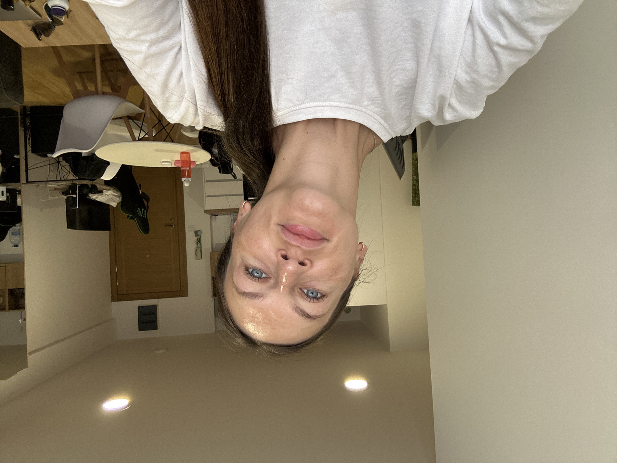 A young woman with blue eyes and long brown hair pulled back in a ponytail takes a selfie in a brightly lit room. She is wearing a white t-shirt. The room appears to be a living space or apartment with light-colored walls and a white ceiling with recessed lighting. In the background, a wooden door is visible, as well as a round table with chairs, a desk with a computer monitor, and shelves. The woman's skin has visible freckles. The scene is located in Podostrog, Montenegro.