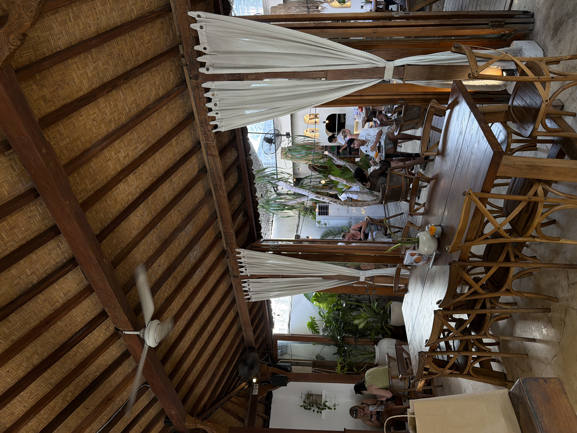 The image captures the interior of a restaurant in Kuta Selatan, Indonesia. The roof is constructed with a textured, woven material supported by dark wooden beams, and a ceiling fan is visible. White curtains are draped between wooden posts, dividing the dining area. Tables and chairs are made of wood and arranged throughout the space, some stacked. People are seated at the tables, engaged in dining. Greenery, including potted plants, is present, adding to the ambiance. The environment appears to be open-air or semi-outdoor, with natural light filtering through. The overall setting is casual and inviting, characteristic of a tropical dining experience.
