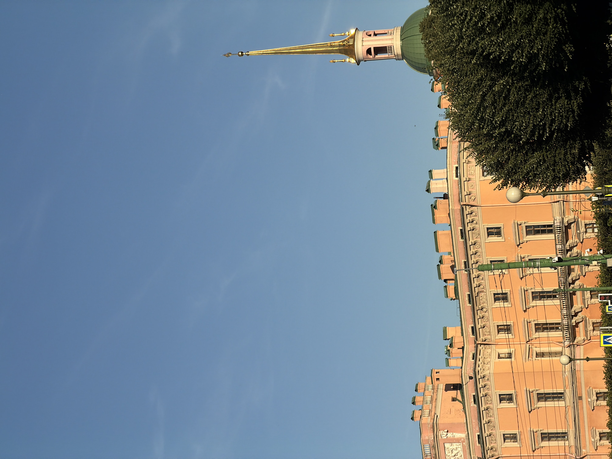 The image shows a section of the Mikhailovsky Castle in Saint Petersburg, Russia, under a clear blue sky. The castle is painted an apricot color and features ornate architectural details, including a decorative cornice and multiple windows. A green-domed structure with a tall, golden spire is visible to the right of the castle, part of the same complex. In the foreground, a dense green tree partially obscures the view of the castle's lower levels. Several lampposts and a section of a street with yellow pedestrian crossing signs are also present. The scene appears to be captured during daylight.