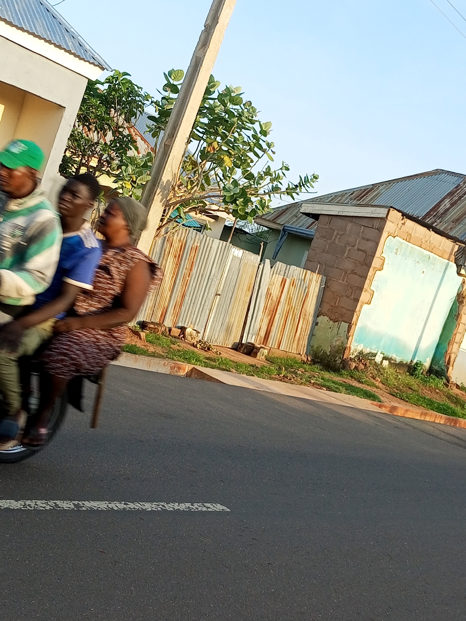 In Gombe, Nigeria, three individuals are riding on a motorcycle. The person in the front is wearing a green cap and a light-colored jacket with green stripes. Beside them, a person in a blue shirt is seated. Behind them, a woman in a patterned dress is the third passenger. They are traveling on a paved road with a white dashed line. To the right of the road, there is a corrugated metal fence and a brick building with peeling blue paint. The scene is illuminated by sunlight, casting shadows on the road. The background consists of a clear sky and vegetation.