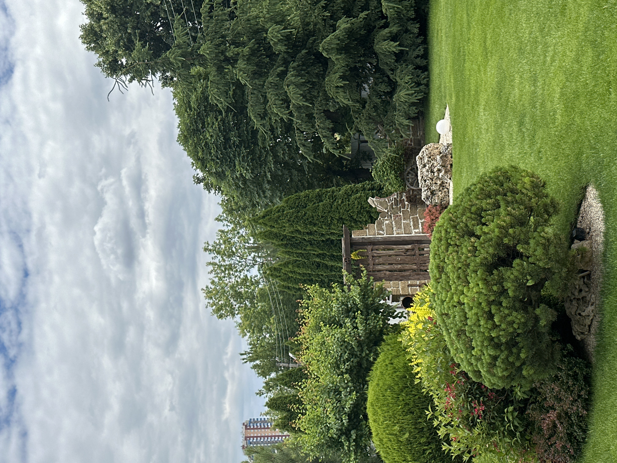 The image depicts a lush, manicured garden in an urban setting. The foreground is dominated by a vibrant green lawn with several meticulously trimmed bushes and trees. In the center, a large, rounded pine-like bush is prominent, set against a backdrop of other green foliage and a rustic wooden gate integrated into a stone wall. Behind the gate, the ruins of a brick structure are visible, adding a touch of historical character. Further back, tall evergreen trees form a dense wall, and a multi-story apartment building stands in the distance, indicating its city location. The sky is overcast with scattered clouds. There are no people or animals visible in the scene. The overall impression is one of a well-maintained and aesthetically pleasing private garden within an urban environment.