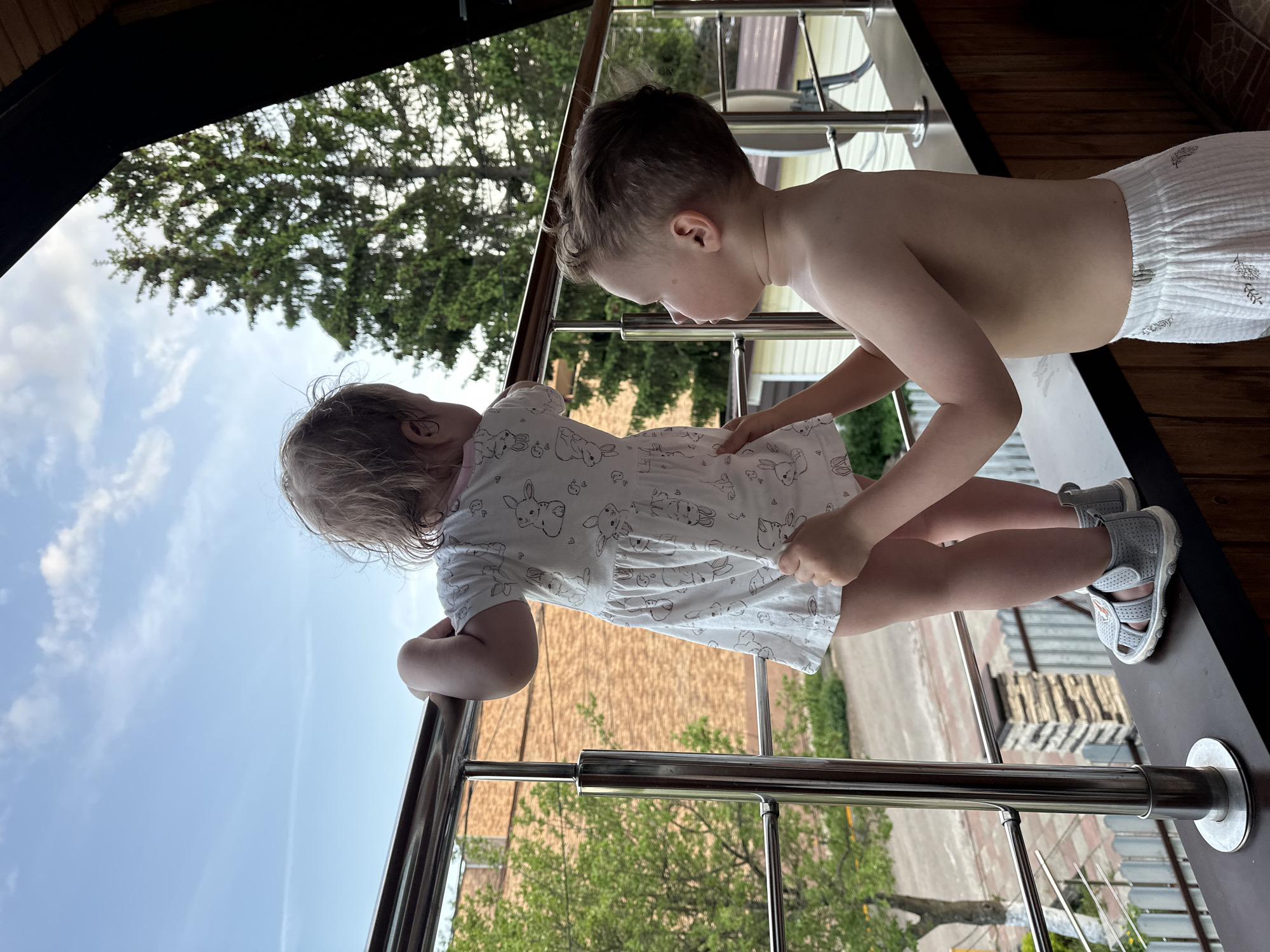 Two young children are on a balcony. A girl with blonde hair wearing a patterned white dress leans against the railing. A boy with brown hair, shirtless and wearing patterned white shorts, stands beside her, his hand touching her dress. They are looking over the edge of the balcony. A tall evergreen tree, blue sky, and a brick building are visible in the background. The location is Nizhny Novgorod, Russia.