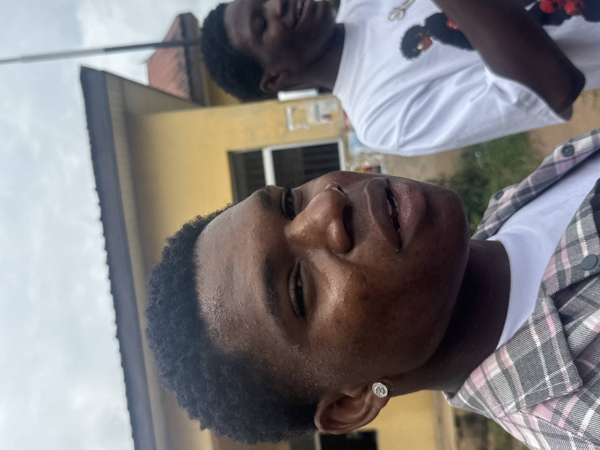 Two young Black males are depicted in this close-up outdoor photograph, which appears to have been taken in Ihiagwa, Nigeria. The primary subject, in the foreground, is facing the camera with his head tilted slightly. He has dark, curly hair and is wearing a checkered shirt over a white t-shirt and a round, silver earring in his left ear. His skin shows some moisture, suggesting warmth or humidity. Behind him, to the right, is another young Black male, also wearing a white t-shirt with a graphic print. He is smiling and looking away from the camera. The background consists of a yellow building with a dark roofline and a partial view of a pole and overcast sky. The overall environment suggests a casual, everyday setting.