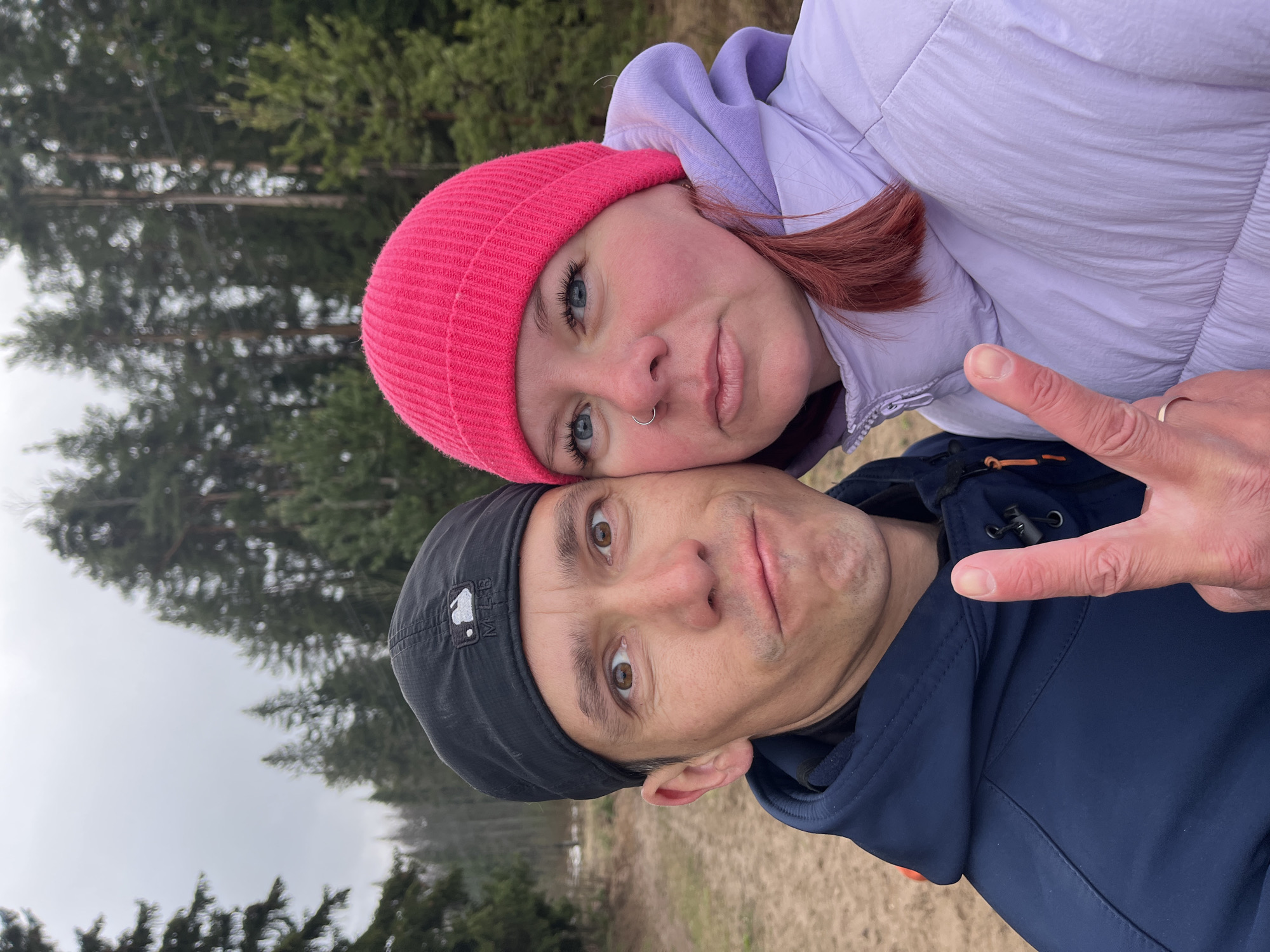 This close-up selfie, captured outdoors in what appears to be a natural setting near Chaikovsky, Russia, features two individuals. The image is oriented vertically, though the subjects are tilted as if the photo was taken in a landscape orientation.
In the foreground, a female subject with reddish-brown hair, blue eyes, and a nose ring (septum piercing) wears a bright pink knit beanie and a light purple hooded jacket. She rests her head gently on the male's head, looking directly at the camera with a calm, slightly smiling expression. Her right hand, adorned with a simple ring on her ring finger, is partially visible on the right edge of the frame.
Beneath her, a male subject with brown eyes wears a black baseball cap featuring a white "MLB" logo and a dark blue hooded jacket. He also looks directly at the camera with a slightly pursed-lip, perhaps playful, expression. His right hand is raised, forming a "V" sign or peace sign with his index and middle fingers.
The background is softly blurred, revealing a dense coniferous forest with tall evergreen trees under an overcast, grey sky. The ground visible behind the subjects appears to be dry, brownish-yellow grass or dirt, suggesting a rural or wilderness location. The attire of both individuals—jackets and a beanie—suggests cool weather. The overall mood is casual and intimate, capturing a shared moment outdoors.