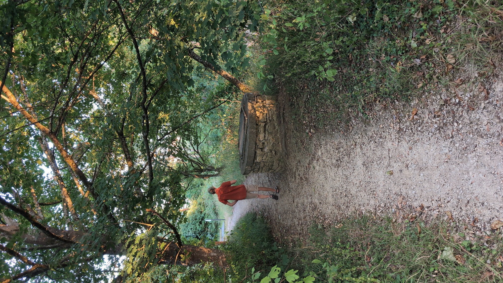 An overhead shot captures a gravel path winding through a sun-dappled forest in Saint-Apollinaire, France. A man wearing an orange shirt and beige shorts walks away from the camera on the path, his back to the viewer. To his right, a stone-built well with a metal grate on top sits beside the path. Lush green foliage and trees with dappled sunlight overhead create a serene, natural environment.