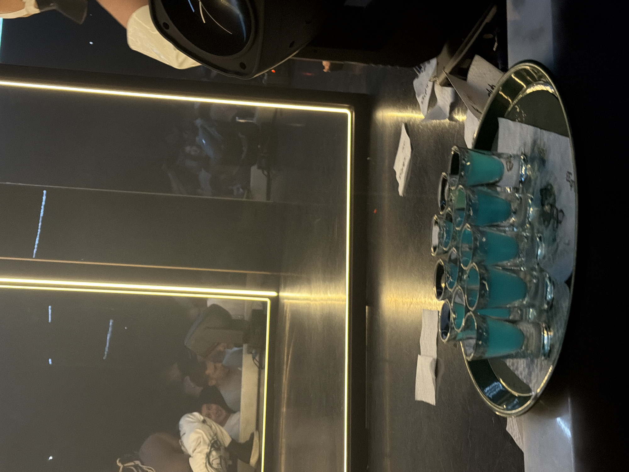 On a dark surface, a silver tray filled with about a dozen shot glasses containing a blue liquid is seen. Small white napkins surround the tray. In the background, reflected in a shiny surface outlined with a gold LED strip, are blurred figures of people. The scene appears to be indoors, possibly at a bar in the city of Sofia, Bulgaria. A black speaker is in the upper part of the image.