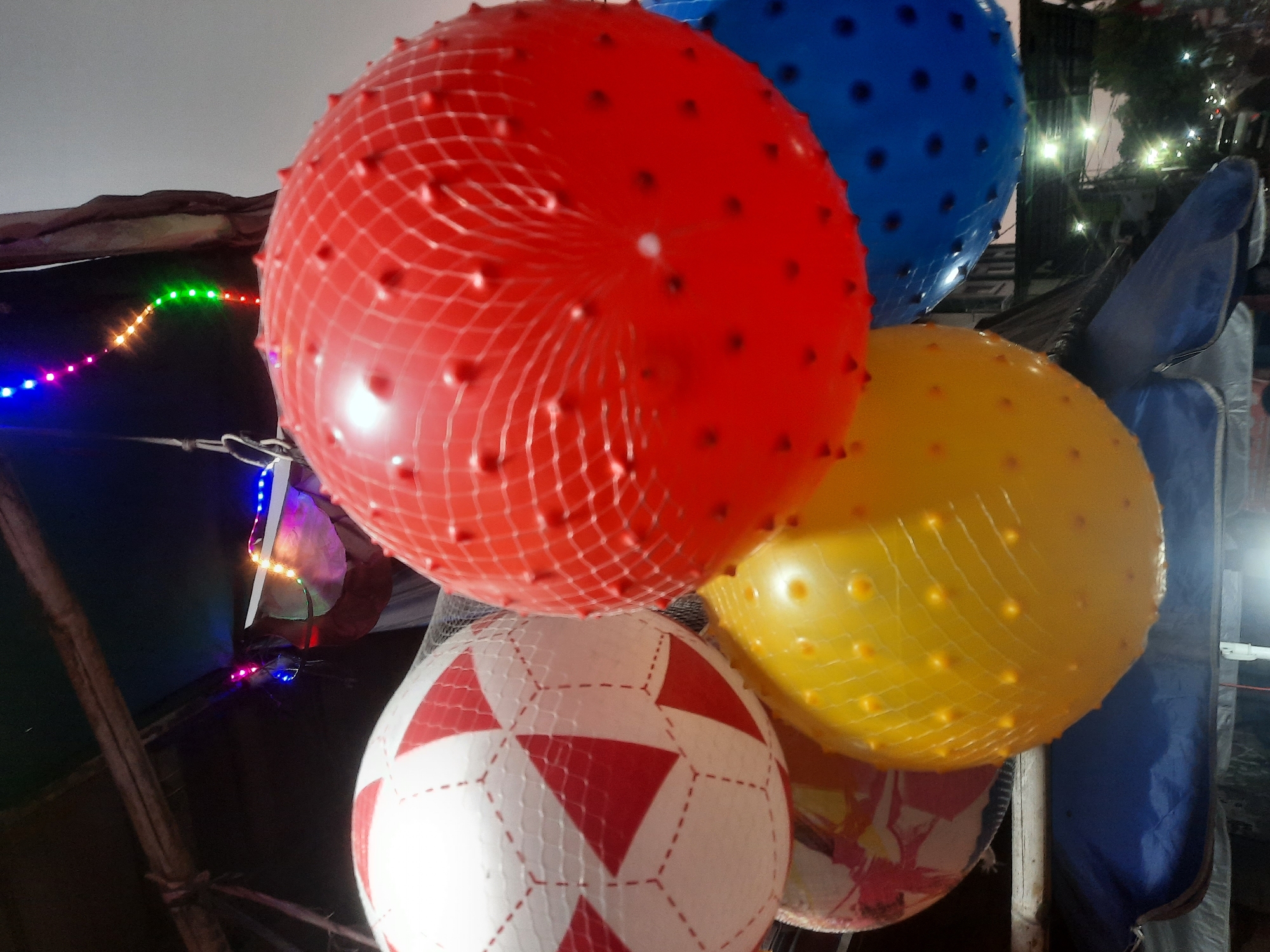 At ground level, outdoors, a collection of various colored inflatable balls are displayed, presumably for sale or as decoration. The balls are suspended from above, possibly from a makeshift stall or structure. Prominently featured are a large red ball with a mesh covering and small raised bumps, a white and red patterned ball resembling a soccer ball, and a bright yellow ball also with a mesh covering and bumps. A portion of a blue ball is visible on the right side. In the background, a string of colorful LED lights is visible, adding a festive element. The scene appears to be set in an urban environment, with blurred lights and structures suggesting a street or market area, possibly in North Jakarta, Indonesia, given the context. The overall impression is that of a lively, possibly nighttime or evening scene, with an emphasis on vibrant, playful objects.