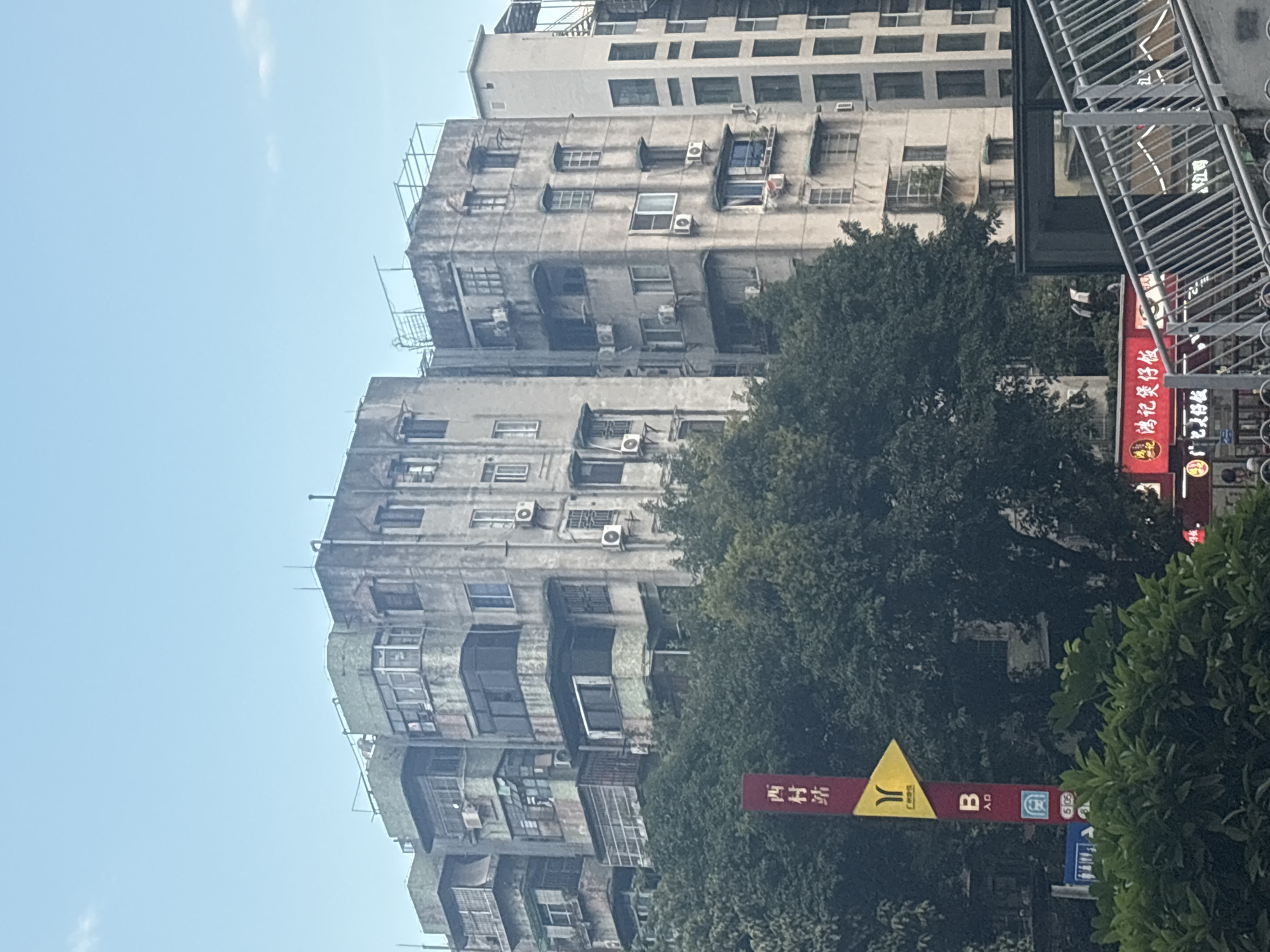 The image depicts an urban scene in Liwan District, China, focusing on several multi-story apartment buildings. The buildings are constructed in a dated architectural style, with weathered concrete facades, visible air conditioning units, and numerous windows. Greenery, primarily dense trees, obscures the lower portions of the buildings. A red sign with Chinese characters and a yellow directional arrow is visible on the left side, indicating a location or establishment. A modern metal railing and staircase are present in the foreground, suggesting a walkway or overpass. The sky above is a clear blue with a few wispy clouds. The overall impression is one of a densely populated urban environment with older residential structures.