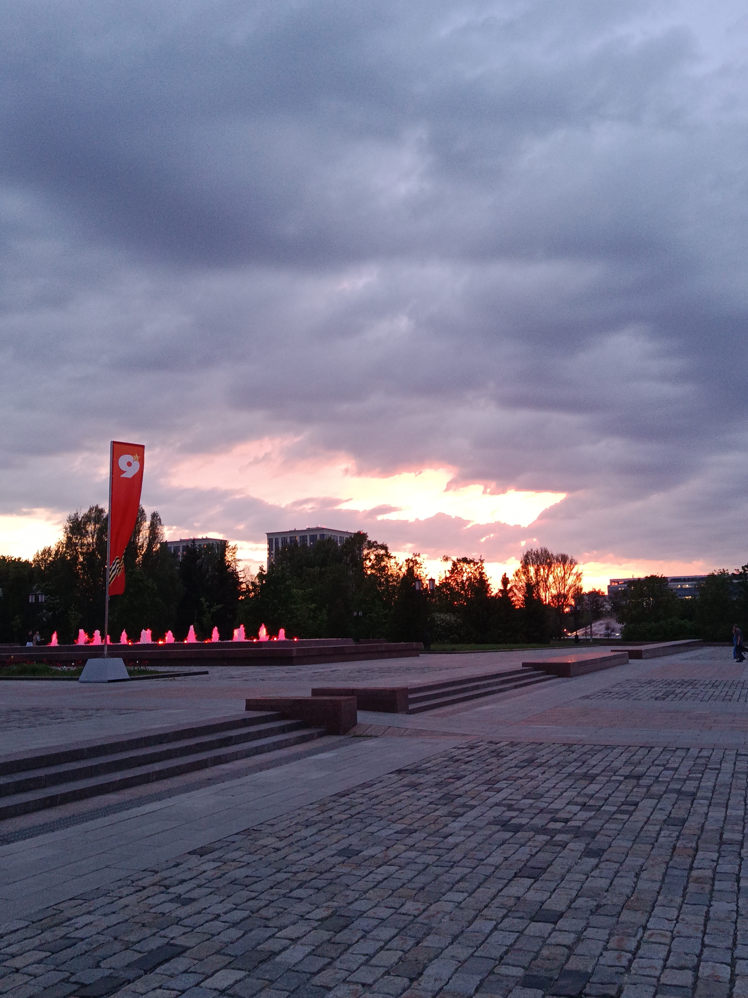 The image shows a sunset scene in Moscow, Russia. The foreground features a paved area with cobblestone patterns and a set of steps leading up to a raised platform. A red flag with a number "9" on it stands prominently on the left, next to a fountain with red-lit water jets. The background includes silhouetted trees and buildings against a sky filled with dark, heavy clouds tinged with orange and pink hues from the setting sun. The overall ambiance suggests a memorial site or public square with a somber yet beautiful atmosphere. A person is walking in the distance on the right side of the image.