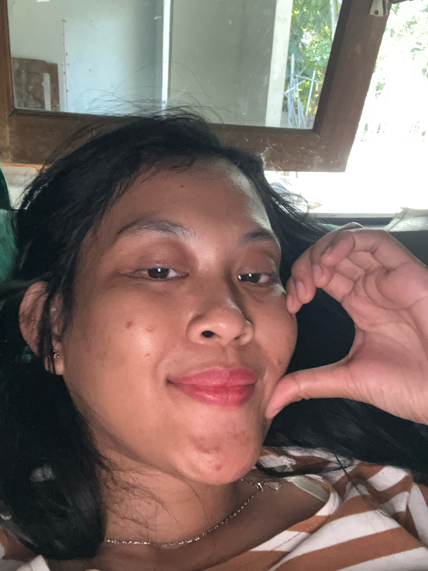 A woman with dark hair and brown skin is taking a selfie in what appears to be a residential setting in Kecamatan Pati, Indonesia. She is looking at the camera and has her right hand near her face, forming a heart shape with her fingers. She is wearing a striped shirt. The background shows a window with trees and foliage visible outside, suggesting an indoor location with natural light. The image is close-up, focusing primarily on the woman's face and upper body.