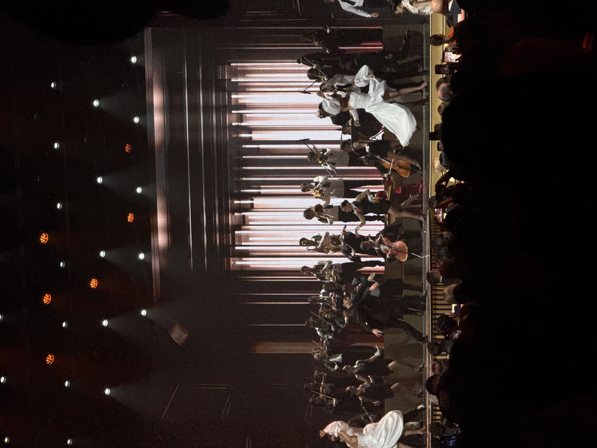 A performance featuring a woman in a white dress and an orchestra in Yerevan, Armenia. The performance is being observed by a crowd, visible to the right. Spotlights illuminate the performance stage. The background features a screen that resembles a set of piano keys.