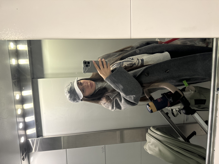 An indoor, full-body mirror selfie depicts a female individual inside an elevator in Akulovo (Gp Kubinka), Russia. The subject, possessing long dark hair, wears a grey newsboy cap, a light grey turtleneck, a grey long double-breasted coat, and a grey graphic sweatshirt. She holds a grey smartphone with a decorative case in her right hand, positioned to capture her reflection. The elevator interior features a reflective surface, metallic paneling on the left, and an overhead section containing four rectangular light fixtures and two ventilation grilles. On the lower left, a light-colored stroller is partially visible, equipped with a dark blue bottle in a holder and a small green attachment. A red handle is partially visible on the lower right.