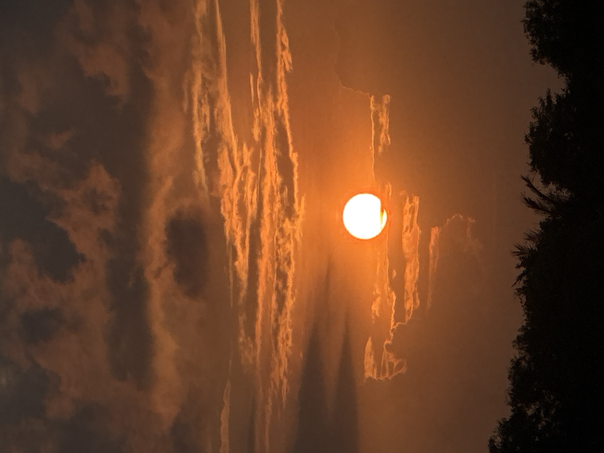 A photograph captures a dramatic sunset over Dayah Leubue, Indonesia. The sky is a vibrant canvas of oranges, reds, and deep browns, with clouds textured by the setting sun's light. A prominent, bright orange sun is positioned in the center of the frame, partially obscured by wispy clouds. Below the sky, a dark silhouette of trees forms the horizon line, suggesting the presence of a forested area. The overall impression is one of natural beauty and the conclusion of a day.