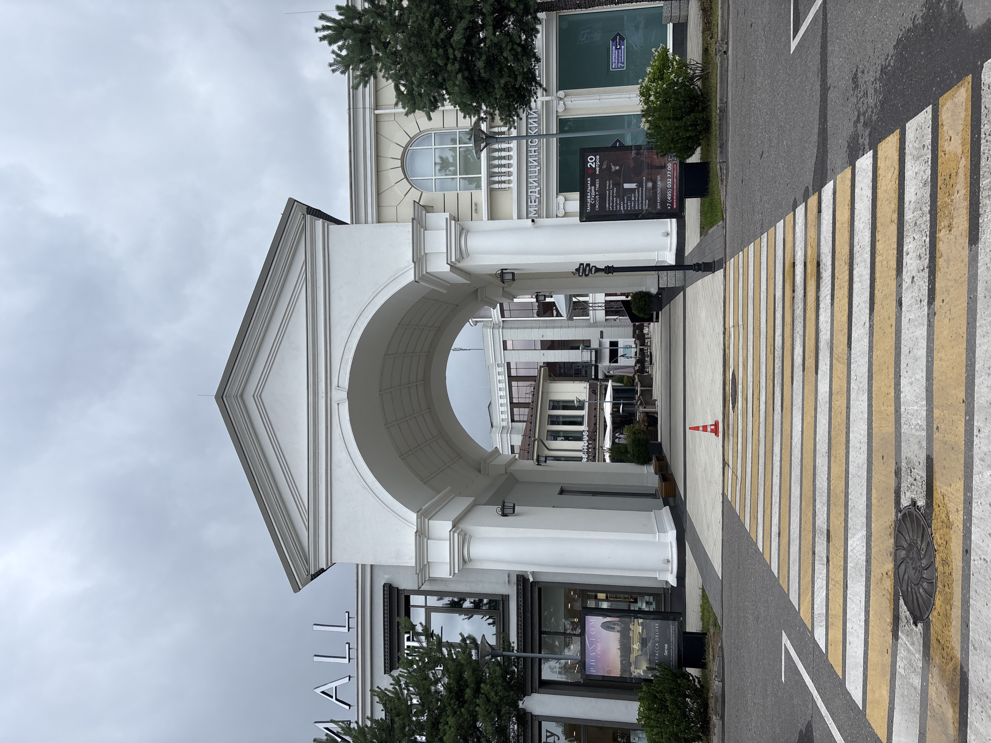 The image shows an outdoor view of an archway leading into a complex, identified as "MILLENNIUM PARK" in Russia. The archway is white with classical architectural elements, supported by large columns. Beyond the archway, a courtyard with more buildings and trees is visible. In the foreground, a pedestrian crossing with yellow and white stripes is prominent on asphalt. A bright orange traffic cone stands on the pedestrian walkway just past the crossing. On either side of the archway are building facades with shops or businesses. On the left, signage reads "MALL" in large silver letters. A poster for "PHANTOM FITNESS" is displayed near the left building entrance. On the right, signage indicates a "MEDICINSKII" center, and another poster promotes dance classes. The sky is overcast and grey. There are no people visible in the immediate scene.