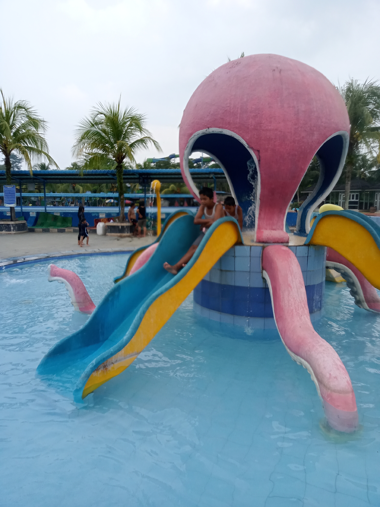 This is a medium-angle, eye-level shot of a water park playground structure in Baru, Indonesia. The main subject is a large, pink, octopus-shaped water slide with blue and yellow slides protruding from its base and dipping into a clear blue swimming pool. Two boys are visible sitting on the edge of the pool at the base of the slides. One boy is wearing a white tank top and the other is wearing a white undershirt. They are facing away from the camera. In the background, there are palm trees, a blue awning structure covering seating areas, and other water slides. Several adults and children are visible in the background, some standing and some sitting. The water in the pool is calm, with ripples around the base of the slides. The sky is overcast.