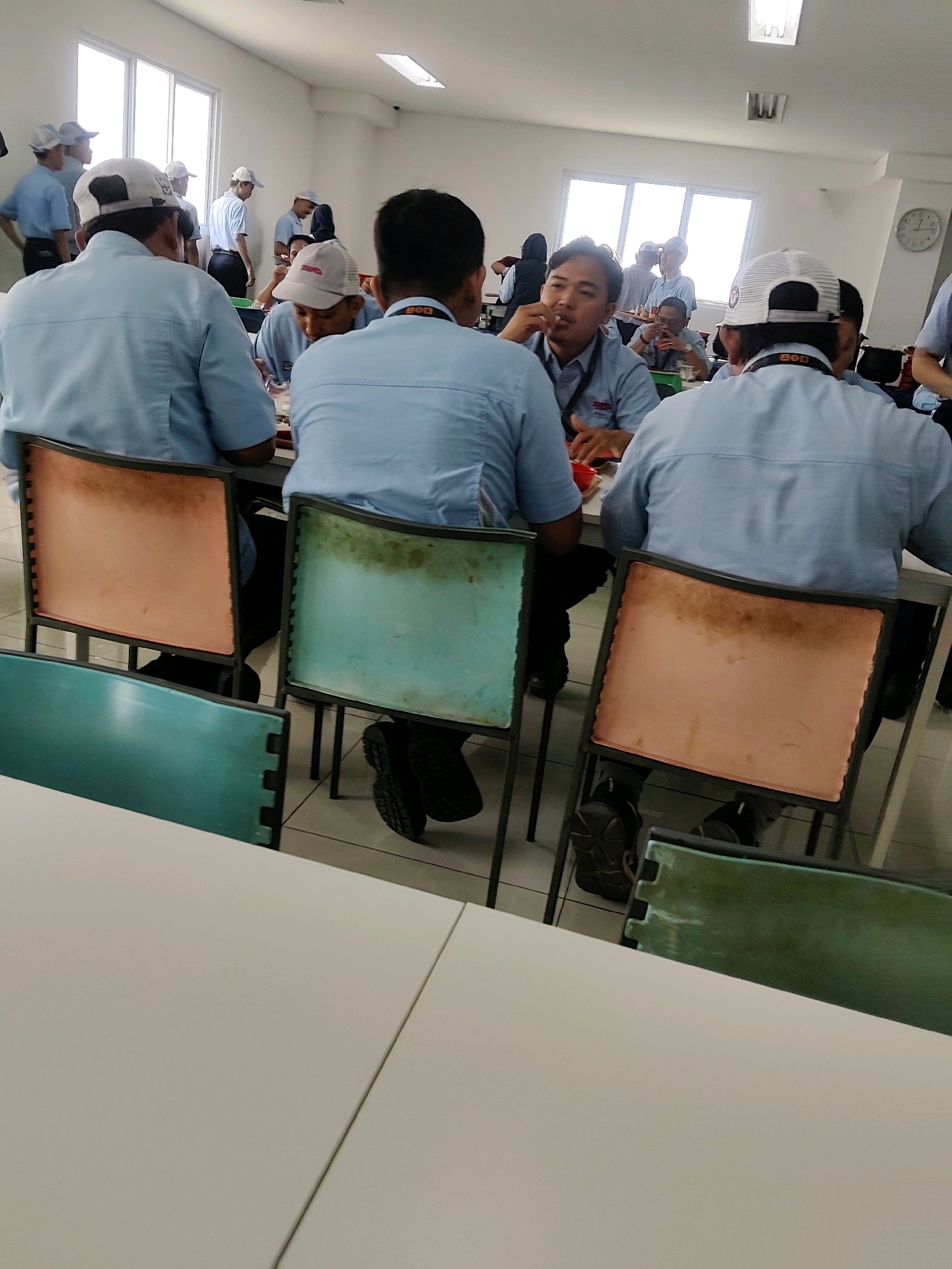 The image shows a group of people in a canteen at a factory, possibly in Mekarmukti, Indonesia. Several individuals wearing light blue shirts and caps are seated at tables, some eating and others talking. One person in the foreground, facing away from the camera, is wearing a light blue shirt and a white cap. Another person, also in a light blue shirt and white cap, is visible from behind in the center right. In the background, more people are standing and milling around. The room appears to be a dining area with tables and chairs. The overall atmosphere suggests a break time or meal service at a workplace.
