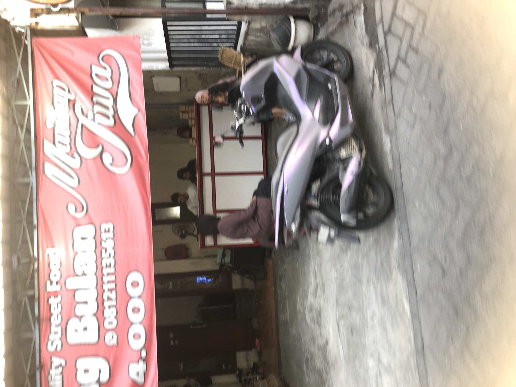 The image depicts an outdoor scene in Tenggulunan, Indonesia, with a focus on a red banner and a motorcycle. The banner displays text in white and pink, with "Quality Street Food" at the top, followed by "g Bulan" and "081211135613", and "4.000" at the bottom. To the right of "g Bulan," stylized text reads "Mantap Jiwa." Behind the banner, a building with an open front reveals several people, some standing and some seated, with what appears to be a counter and boxes of goods. A person in a dark purple garment is seated on a bench in front of the building. A light purple and gray scooter is parked in the foreground, facing left. The ground is paved with concrete and some stones. The scene appears to be a street-side food stall or shop.
