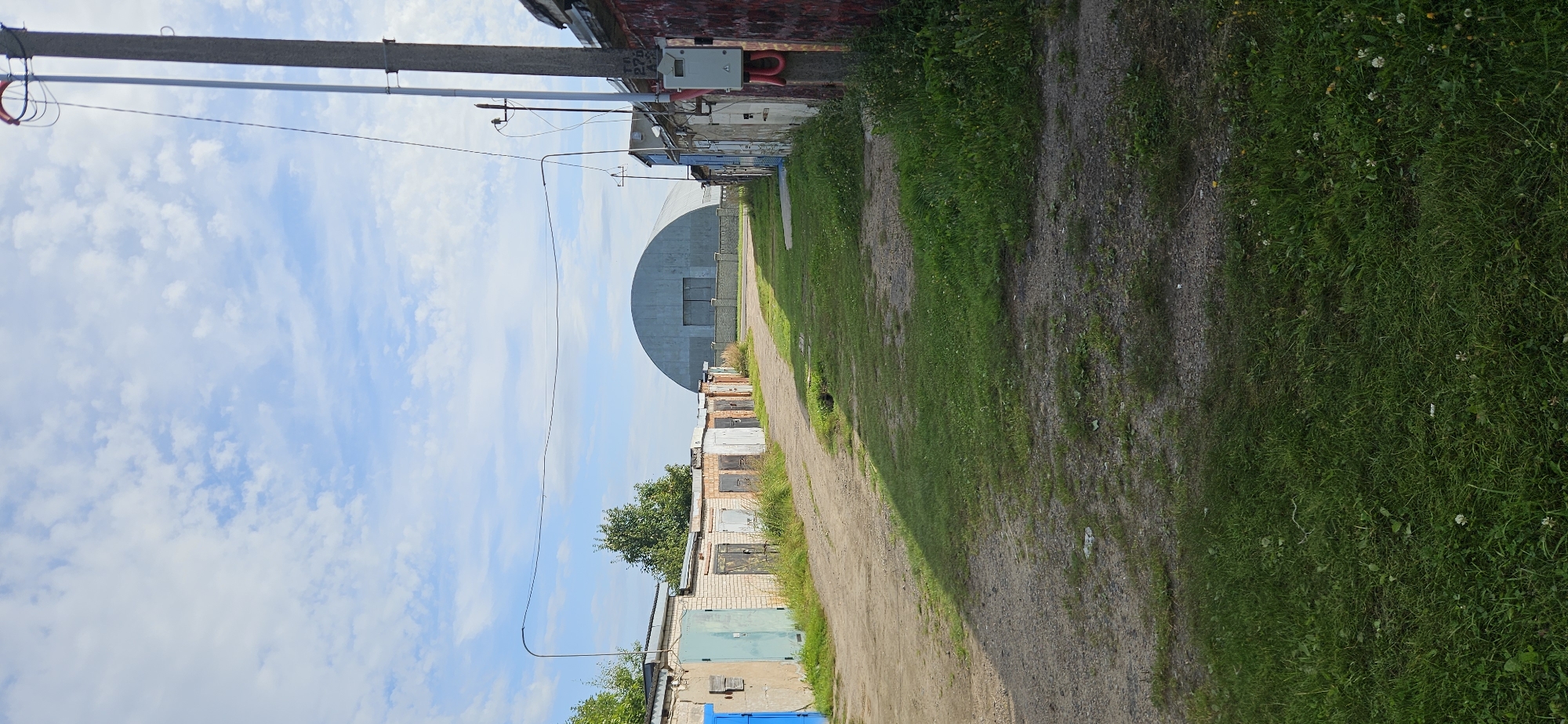 A vertical shot captures a dirt and grass pathway in what appears to be a garage cooperative. On the left, a row of brick garages with various colored doors lines the path. A large, curved, grey industrial building with a large door is visible in the distance, centered between the rows of garages. On the right, another row of garages is partially visible, with a utility pole in the foreground adorned with electrical equipment. The sky is blue with scattered white clouds. The scene is bathed in daylight, casting shadows across the pathway and grass. There are no people or animals present in the image.