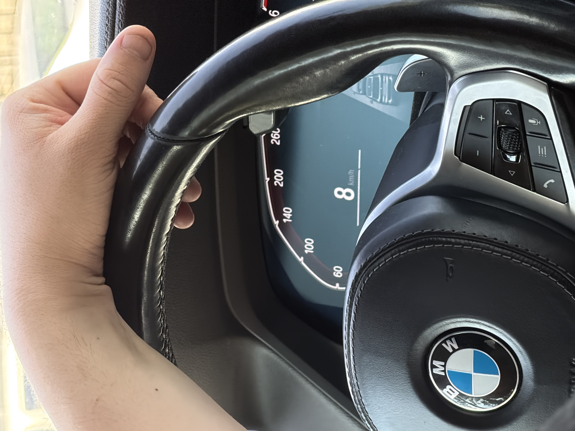 A close-up view inside a BMW vehicle is presented, with a hand visible on the left side of the steering wheel. The dashboard and BMW logo on the steering wheel are clearly visible. The speedometer reads 8 km/h. The location cannot be ascertained from the image.