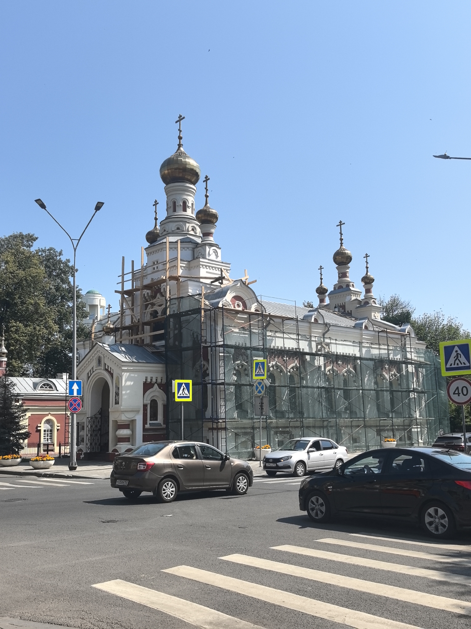 The image shows an Orthodox church in Nizhny Novgorod, Russia, undergoing renovation. The church has a white facade with red accents and several golden domes topped with crosses. Scaffolding and green netting are visible, indicating ongoing construction work. Several cars are present on the road, including a brown sedan in the foreground and a silver sedan approaching a crosswalk. Traffic signs, including a pedestrian crossing sign and a speed limit sign of 40, are visible on the right side of the image. The sky is clear and blue, suggesting a sunny day. The church is situated in an urban environment, with trees and other buildings visible in the background.