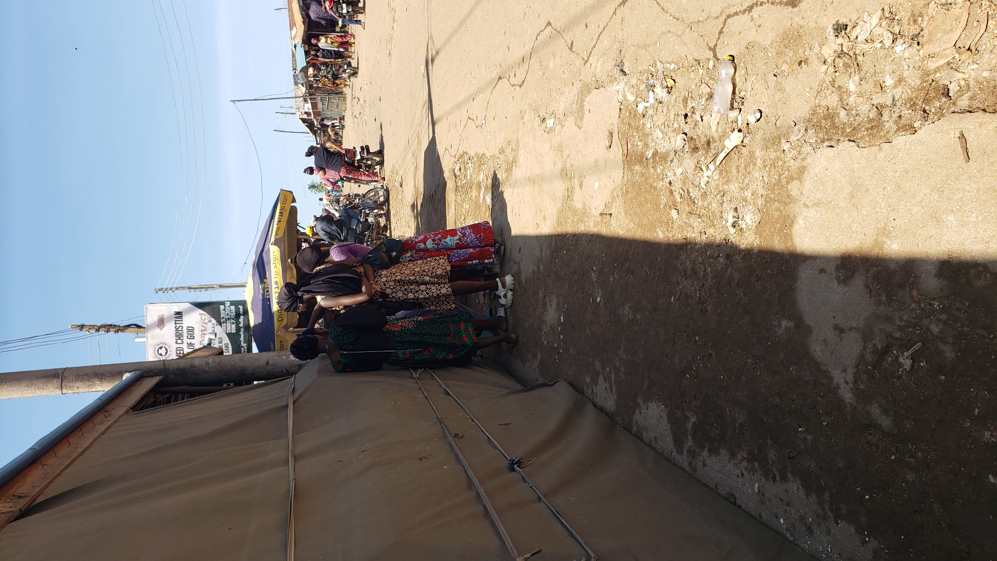 The image depicts an outdoor street scene under a clear blue sky in Sabon Tasha, Nigeria. A large, temporary-looking structure covered in light brown canvas or tarp material occupies the left foreground, casting a long, dark shadow diagonally across the uneven, dusty ground. This ground is a mix of dirt and cracked concrete, with scattered debris including plastic bottles and other litter.
Four women are clustered together in the mid-left, their backs mostly to the camera, standing adjacent to the temporary structure. From left to right, one woman wears a dark top with a green and white patterned skirt; the next wears a dark top and a leopard-print skirt; another wears a dark top and a long, red, white, and blue patterned skirt. They appear to be engaged in conversation or waiting.
A tall utility pole stands slightly right of center, affixed with a rectangular sign that reads "RED CHRISTIAN OF GOD," "David," and partially visible service times "OURS: 5.30PM" and "THURS: 5.30PM" below "SABO," confirming the location. Behind the women, a yellow and blue striped canopy or umbrella is visible, displaying text that includes "Dial *671# to start."
Further down the unpaved road, in the mid-ground, several individuals are visible. Two men are seated on motorcycles, one wearing a yellow helmet, the other a darker one. Another person is near a red motorcycle. The background shows an array of informal structures, likely market stalls or small buildings, characterized by corrugated metal roofs and walls, extending along the dusty street where more indistinct figures are present. The scene is illuminated by strong sunlight, indicating a bright day.