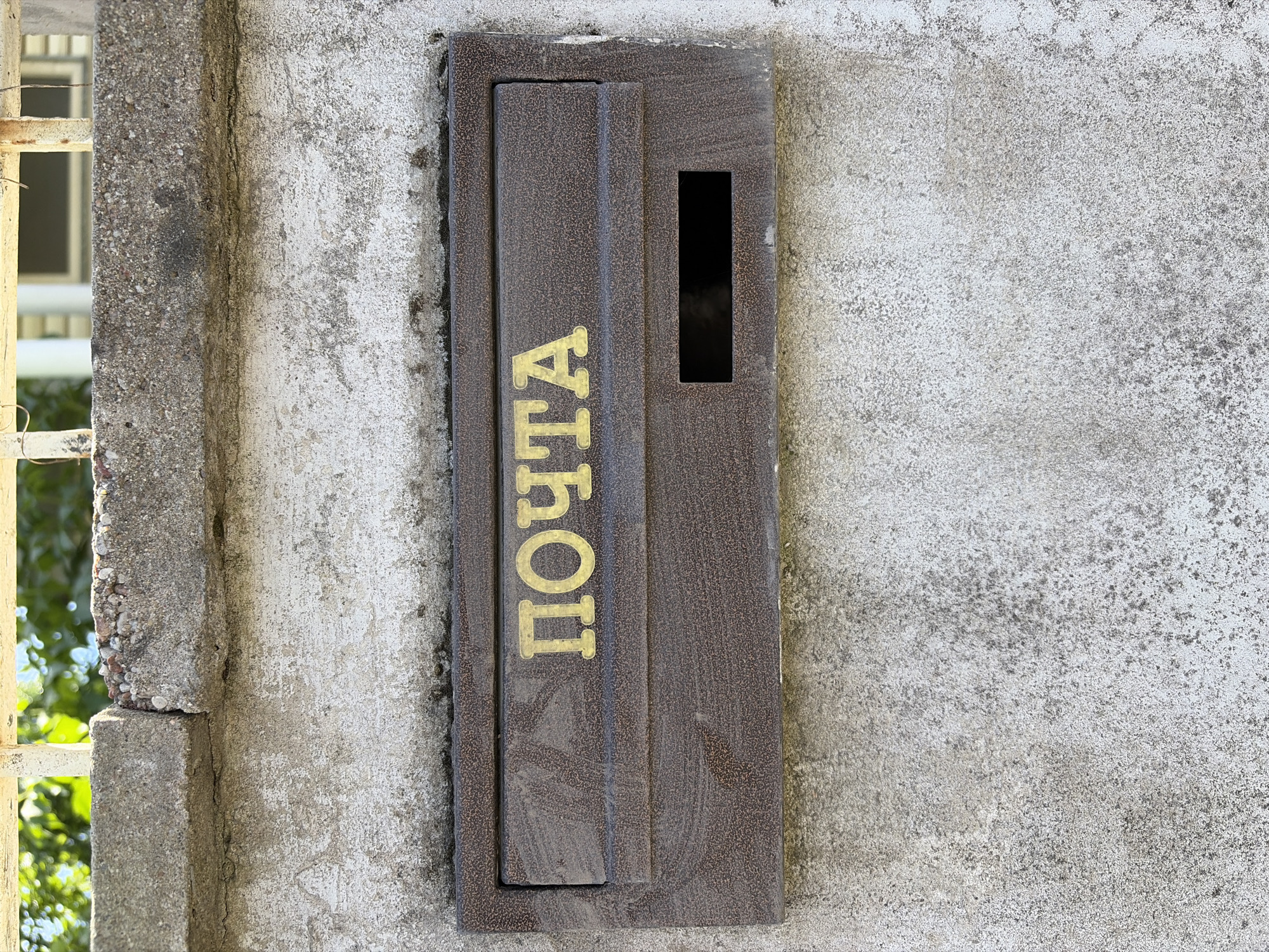 A close-up, frontal view shows a weathered, brown metal mailbox mounted on a textured concrete wall. The mailbox has a rectangular slot for mail and the word "ПОЧТА" (which translates to "POSTA" or "MAIL") is emblazoned in gold lettering above the slot. The wall is rough and appears to have areas of discoloration and staining. Above the mailbox, a concrete ledge runs horizontally, with a metal railing or fence visible at the very top of the image, silhouetted against a blurred background of greenery and what appears to be part of a building. The scene suggests an outdoor setting, likely a residential or public area.