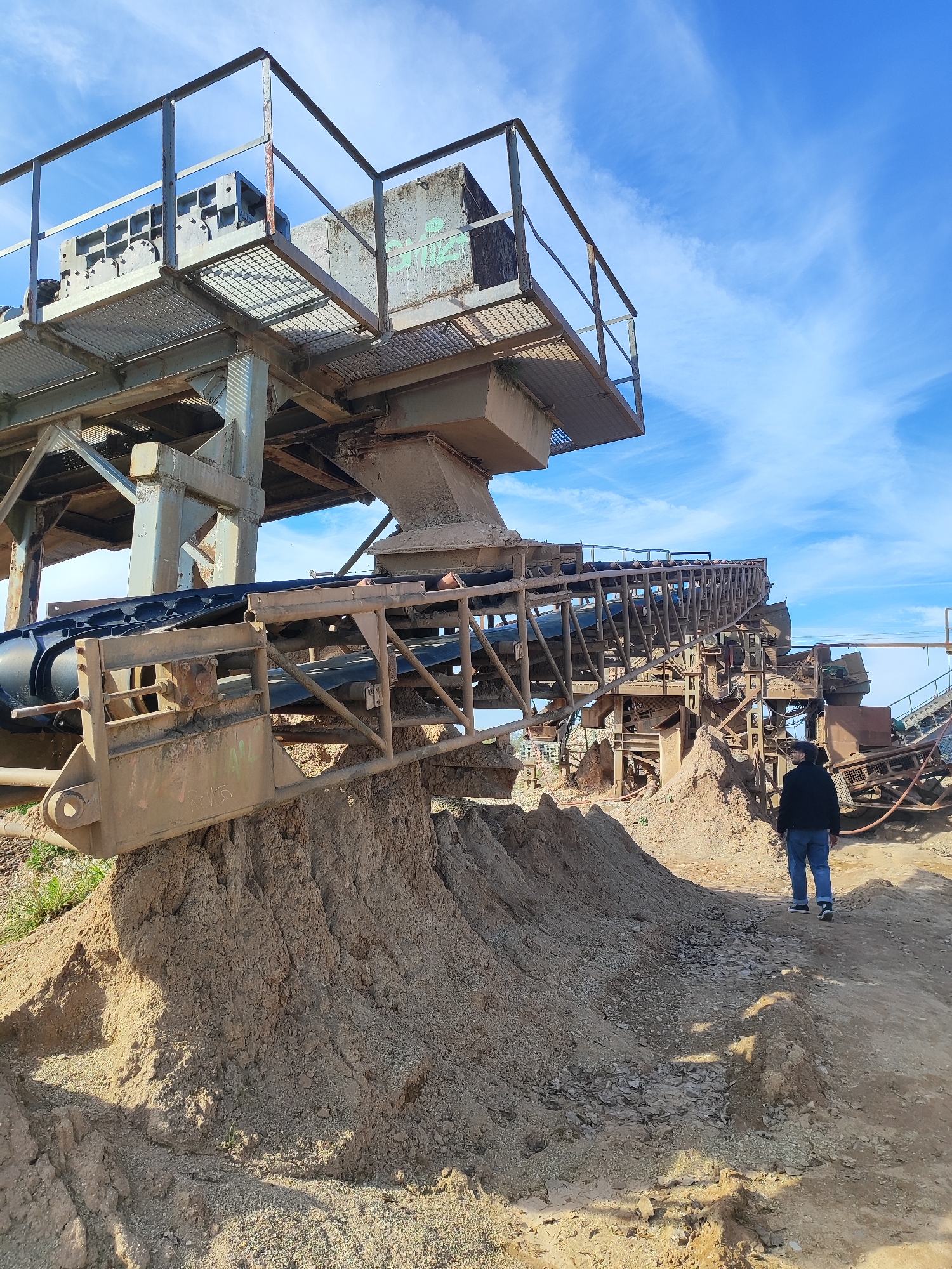 The image shows a man walking on a dirt path in an industrial setting. To the left is a large, tan-colored machine with metal framework and conveyor belts, sitting atop a pile of dirt. The sky is blue with scattered clouds. The location is Unknown, Unknown.