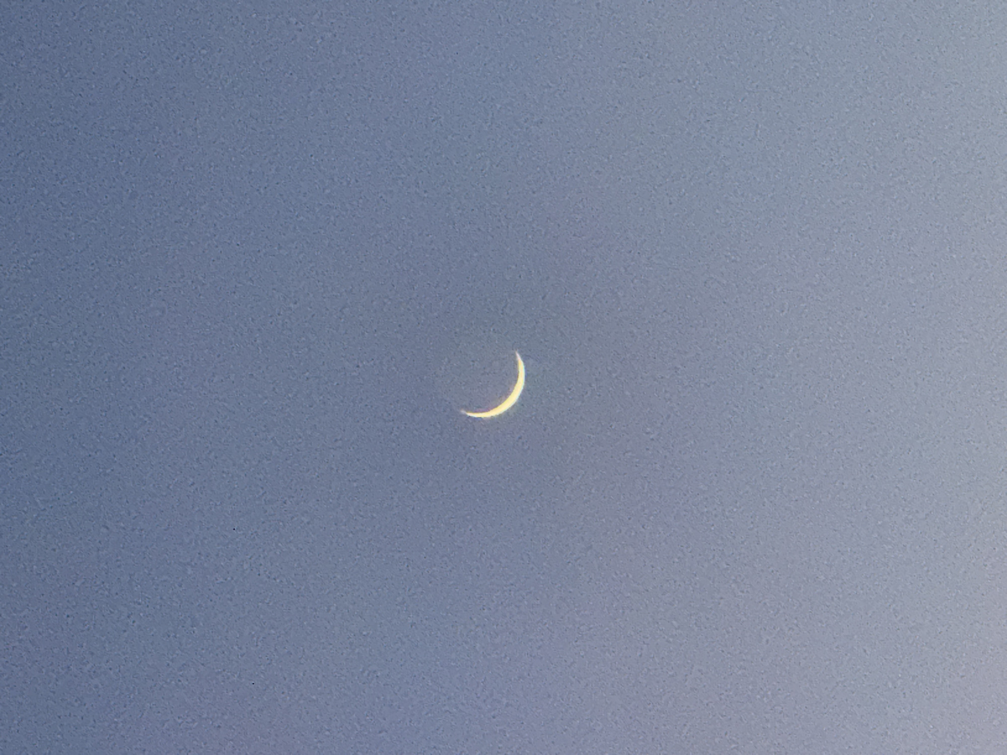 The image shows a crescent moon in a clear, light blue sky. The location, Kuta Selatan, Indonesia, is inferred as metadata is not provided. There are no people, subjects, objects, or actions depicted besides the moon's presence in the sky. The environment is a daytime or twilight sky based on the color.