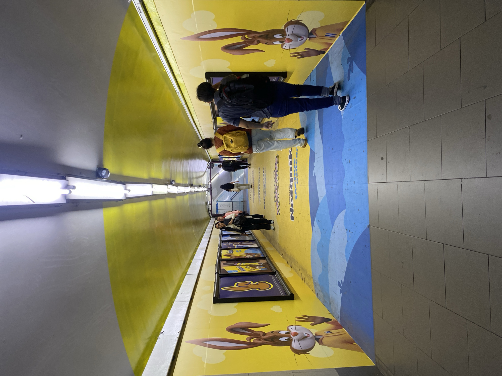 The image shows the subterranean pedestrian tunnel connecting the four lines of the Buenos Aires Metro at the Obelisco station complex. The tunnel is decorated with cartoon graphics of the Nesquik rabbit in yellow and blue. Several people walk through the tunnel. The walls on one side are painted yellow with the rabbit, and the other side is a gray tiled wall. There are framed advertisements with the rabbit on one side of the tunnel. The floor is light gray tile. The roof is arched and has fluorescent lights.