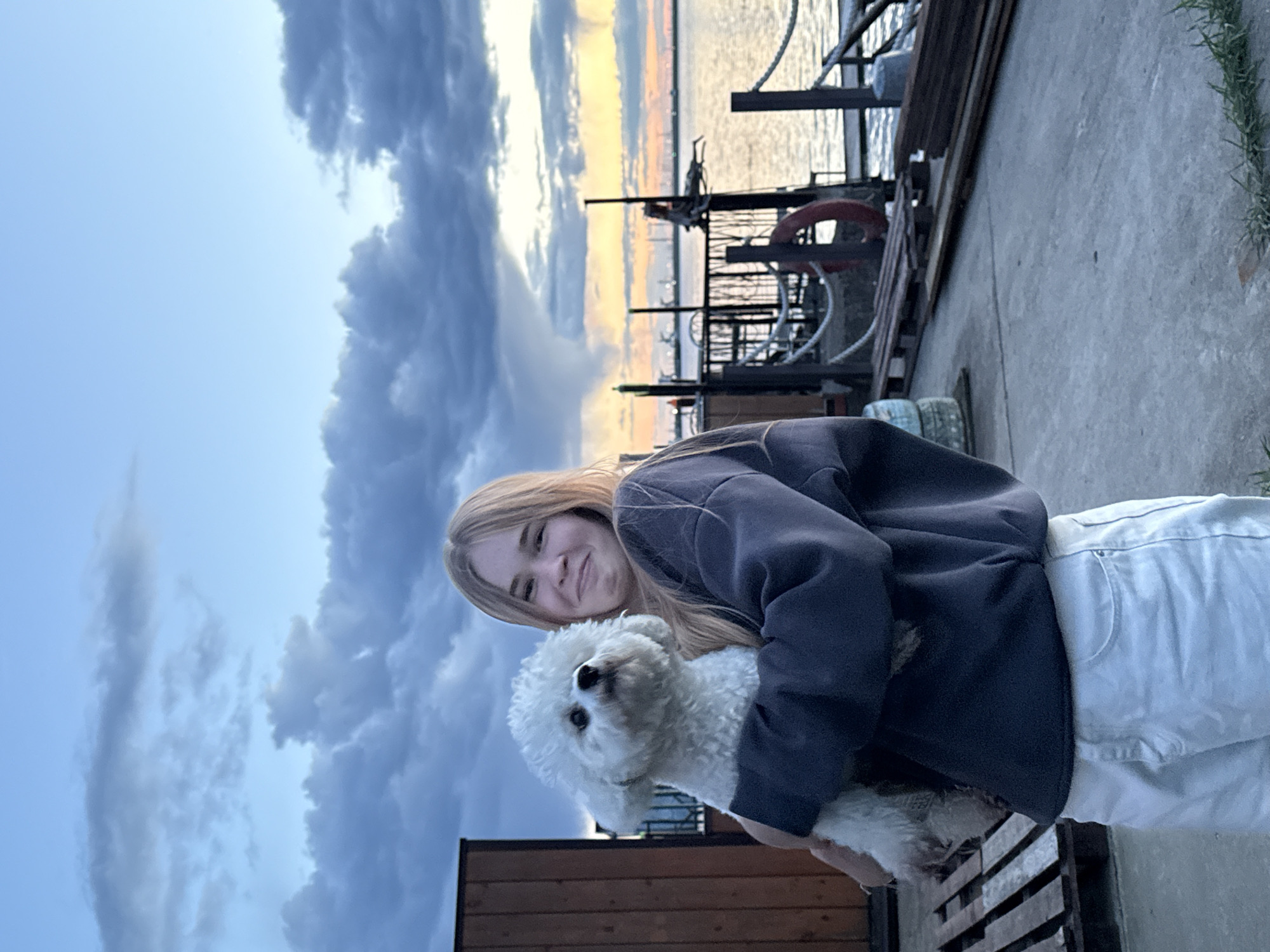 A young woman with blonde hair is holding a white dog in her arms. They are standing on a concrete pier with a metal railing behind them. The setting sun is reflected on the water, and there are dark, dramatic clouds in the sky. The location is Chaikovsky, Russia.