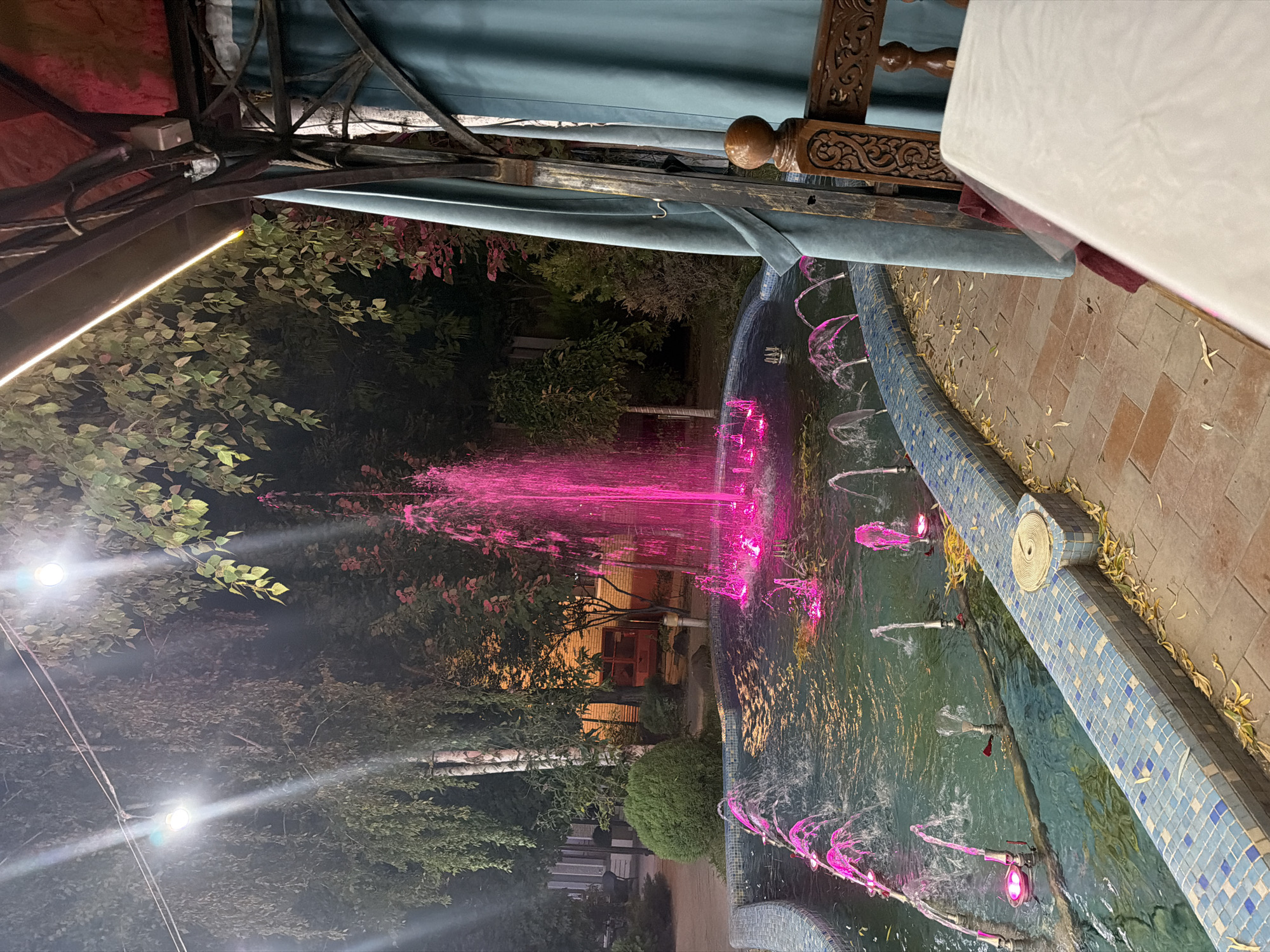 An outdoor fountain with bright pink water jets is illuminated at night. The fountain is surrounded by a tiled border and a paved walkway. Lush green trees and foliage are visible in the background, along with some outdoor lighting fixtures. To the right, a portion of a covered seating area with turquoise curtains and a wooden chair with carved details can be seen, along with a white-covered table. The location is identified as Chinarkent, Uzbekistan.