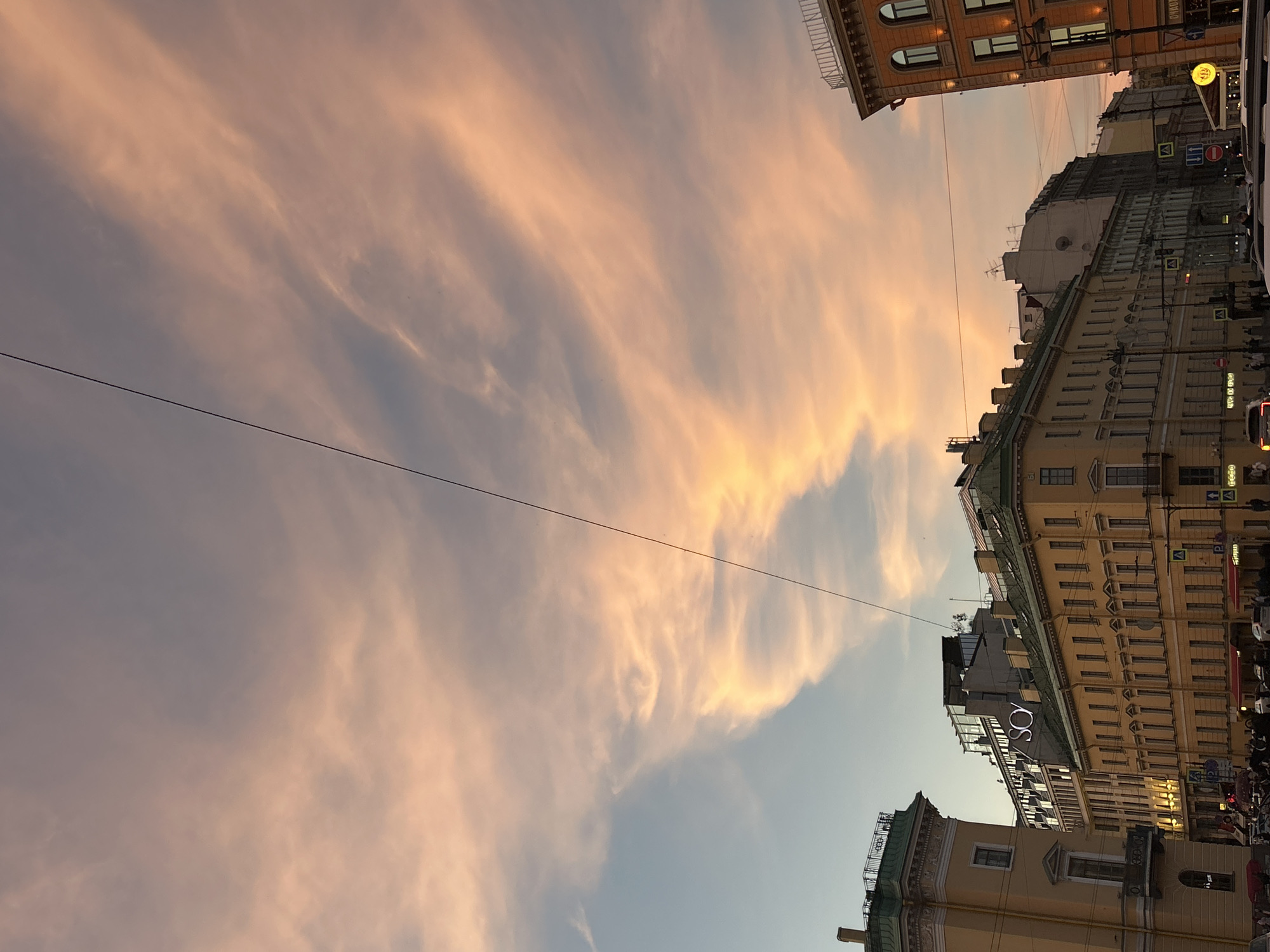 This image captures a street view of buildings with a pink and orange hued sky with wispy clouds at dusk. The sky takes up a significant portion of the frame, with a thin black wire diagonally crossing it. The buildings are multi-story and appear to be in an urban setting, with architectural details like windows, balconies, and rooftops visible. One building prominently displays the illuminated sign "SOY." Pedestrians and vehicles are present on the street below, suggesting a busy city environment. The lighting and colors indicate the time of day is likely around sunset or sunrise.