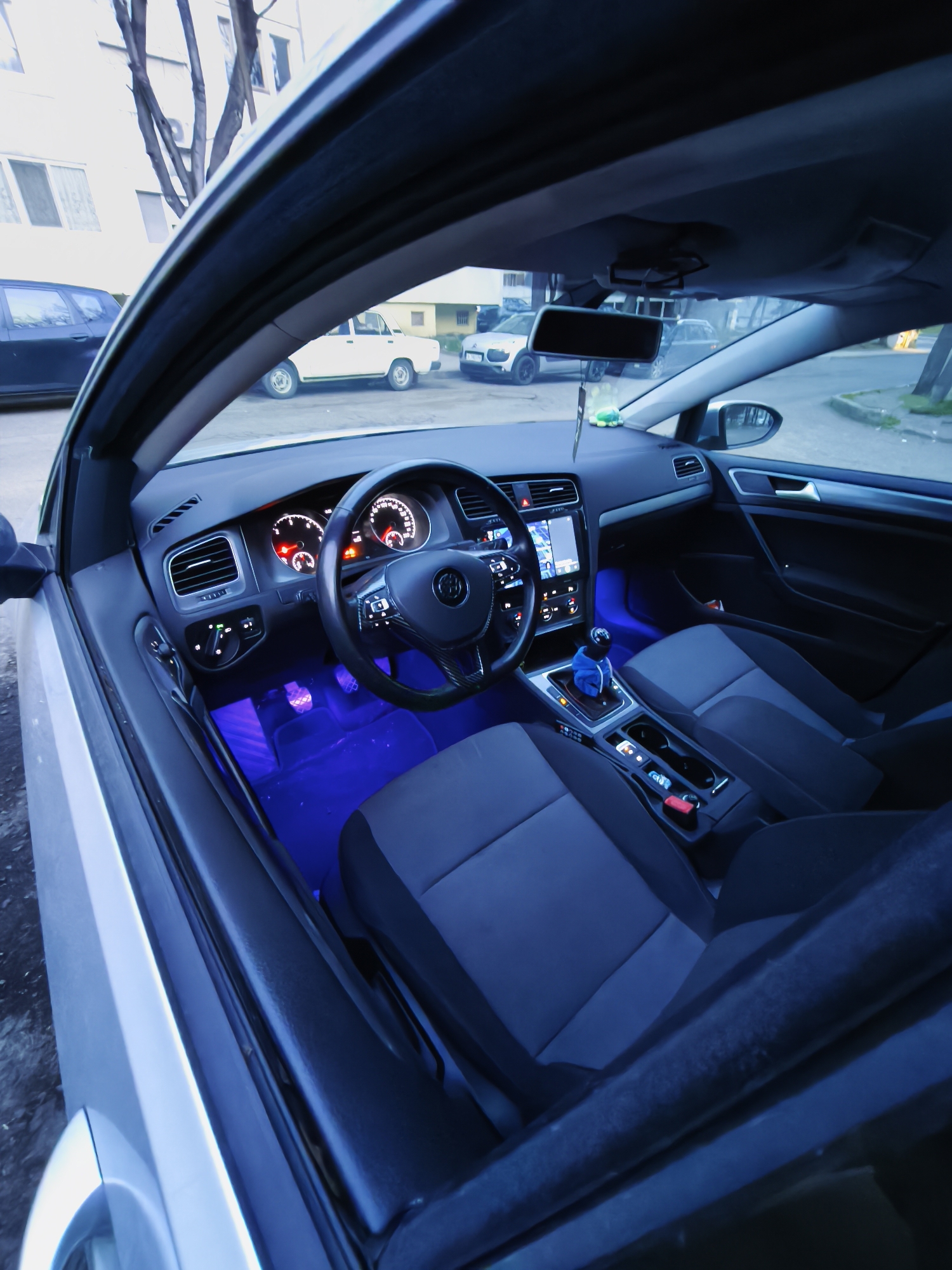 The image shows the interior of a Volkswagen Golf, likely a late-model version (MK7 or MK7.5) given the dashboard layout and infotainment screen. The interior features aftermarket blue ambient lighting. The car has a manual transmission, identifiable by the stick shift in the center console. Outside the car, through the open driver-side door, there are several other parked cars. A Lada and another hatchback are visible. The location is likely Burgas, Bulgaria, as suggested. The environment appears to be a residential or mixed-use area. The lighting suggests dusk or early evening.