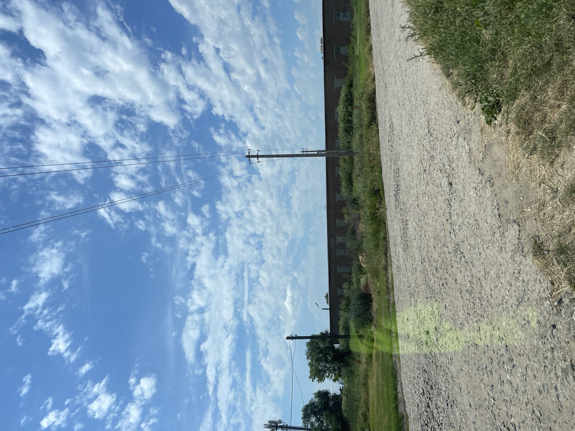 The image captures a daytime scene on a gravel road in Bezopasnoye, Russia. In the foreground, the textured surface of the road is visible, with patches of dry grass and green vegetation growing along the edges. A faint, vertical green light reflection is present on the road's surface, possibly from a vehicle.
In the middle ground, a long, low building constructed of brick is situated behind a line of green shrubs and grass. The building appears to have numerous small, dark windows or openings evenly spaced along its facade. Several utility poles with electrical wires strung between them are prominent features, one standing tall in the center of the frame and another to the left, near a large cellular tower. Trees, some with dense foliage, are visible to the left of the building and poles.
The background is dominated by a bright blue sky filled with scattered, puffy white clouds. The overall impression is one of an industrial or semi-rural area under a clear sky.