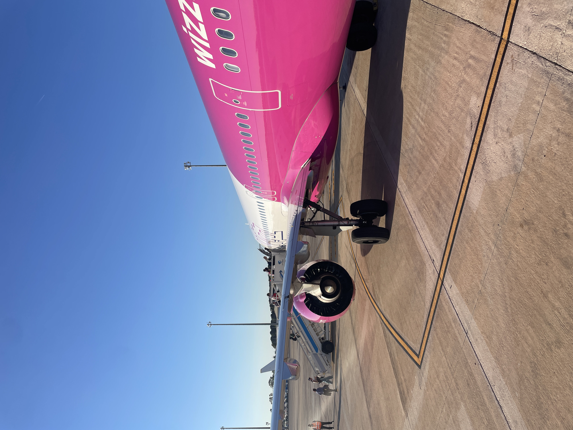 The image depicts a Wizz Air aircraft on the tarmac at what appears to be an airport. The fuselage is predominantly pink with the airline's name clearly visible. An engine is mounted under the wing. Passengers are boarding the plane via a mobile staircase. The airport setting is indicated by the paved surface with painted taxi lines, lighting poles, and other aircraft visible in the background. Based on the image content and the description provided, it is likely that this photograph was taken at Valencia Airport (VLC) which is located in the city of Manises, Spain. There are people walking nearby and passengers are embarking on the plane. The sky is clear and blue.
