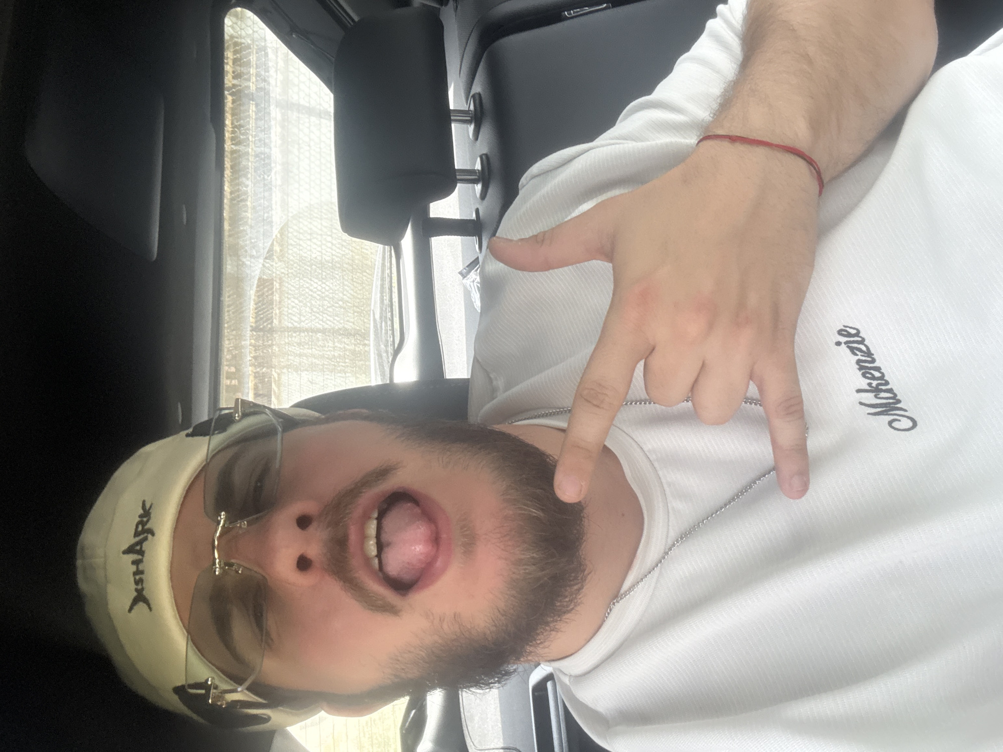 A bearded man wearing a white shirt with "Mckenzie" embroidered on it, a light-colored cap with "JSHARK" printed on it, and glasses, is in a vehicle. He is making a hand gesture with his tongue sticking out. A red string bracelet is on his wrist. The background shows the interior of a vehicle, including a window and headrest. The location is unknown, and the text does not suggest the city of Teteven, Bulgaria.