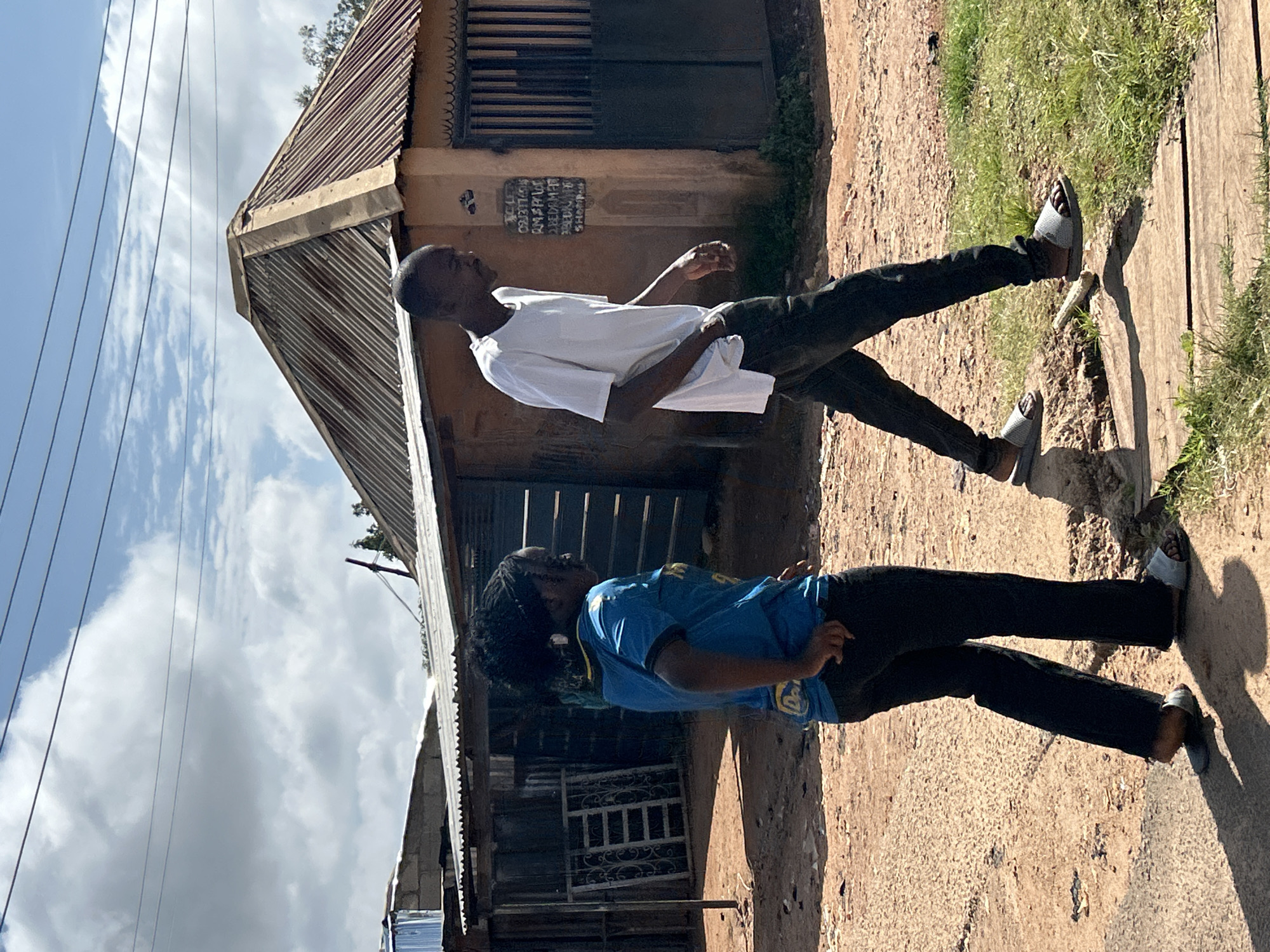 In an outdoor, urban environment in Jos, Nigeria, two individuals walk on a dirt path. The person on the left, facing away from the camera, is a woman wearing a bright blue t-shirt with yellow trim and black pants. She has braided dark hair and is wearing light-colored sandals. The person on the right is a man, also facing away, wearing a white t-shirt and dark pants, and appears to be wearing light-colored sandals.
Behind the individuals are structures with corrugated metal roofs. The building on the right has tan walls with a dark, barred doorway. A sign with text is affixed to the wall next to the doorway. To the left, there is a smaller structure with a silver corrugated metal roof and what appears to be a metal grate leaning against its side. The ground is a mix of dirt and patches of grass, with some wooden planks forming a walkway. The sky above is partly cloudy with blue sky visible.