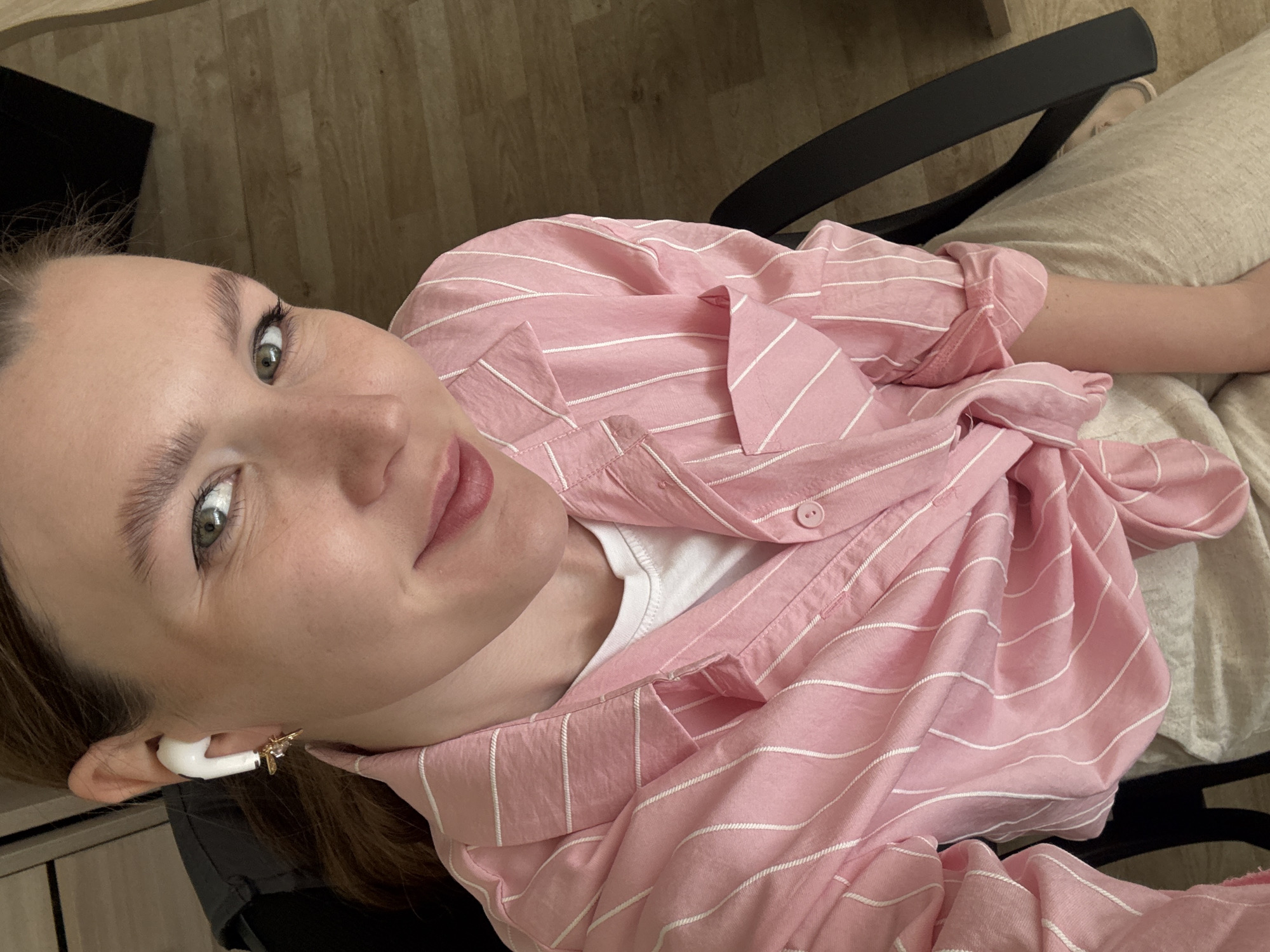 A woman is sitting in a chair. She has light skin, green eyes, and brown hair. She is wearing a pink and white striped shirt over a white t-shirt, and light beige pants. She has a white wireless earbud in her left ear, adorned with a gold and clear gem embellishment. The chair has a black back. The background is a light brown wooden floor. The location is not evident in the image to confirm it is in Izhevsk, Russia.