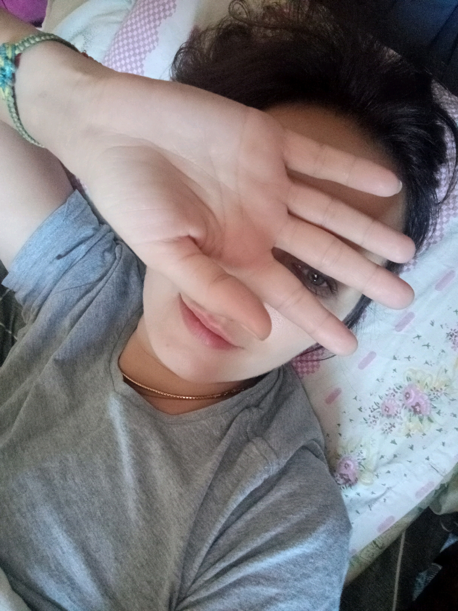 A person is lying on their back on a bed, covered by a floral-patterned blanket and a pink pillow. They are wearing a gray t-shirt and a thin gold necklace. Their left arm is raised, and their hand is covering their face, obscuring their eyes and forehead. A beaded bracelet with blue and yellow elements is visible on their left wrist. The person's dark hair is spread out around their head. The overall impression is one of relaxation or possibly an attempt to avoid being photographed. The location is not specified in the image.