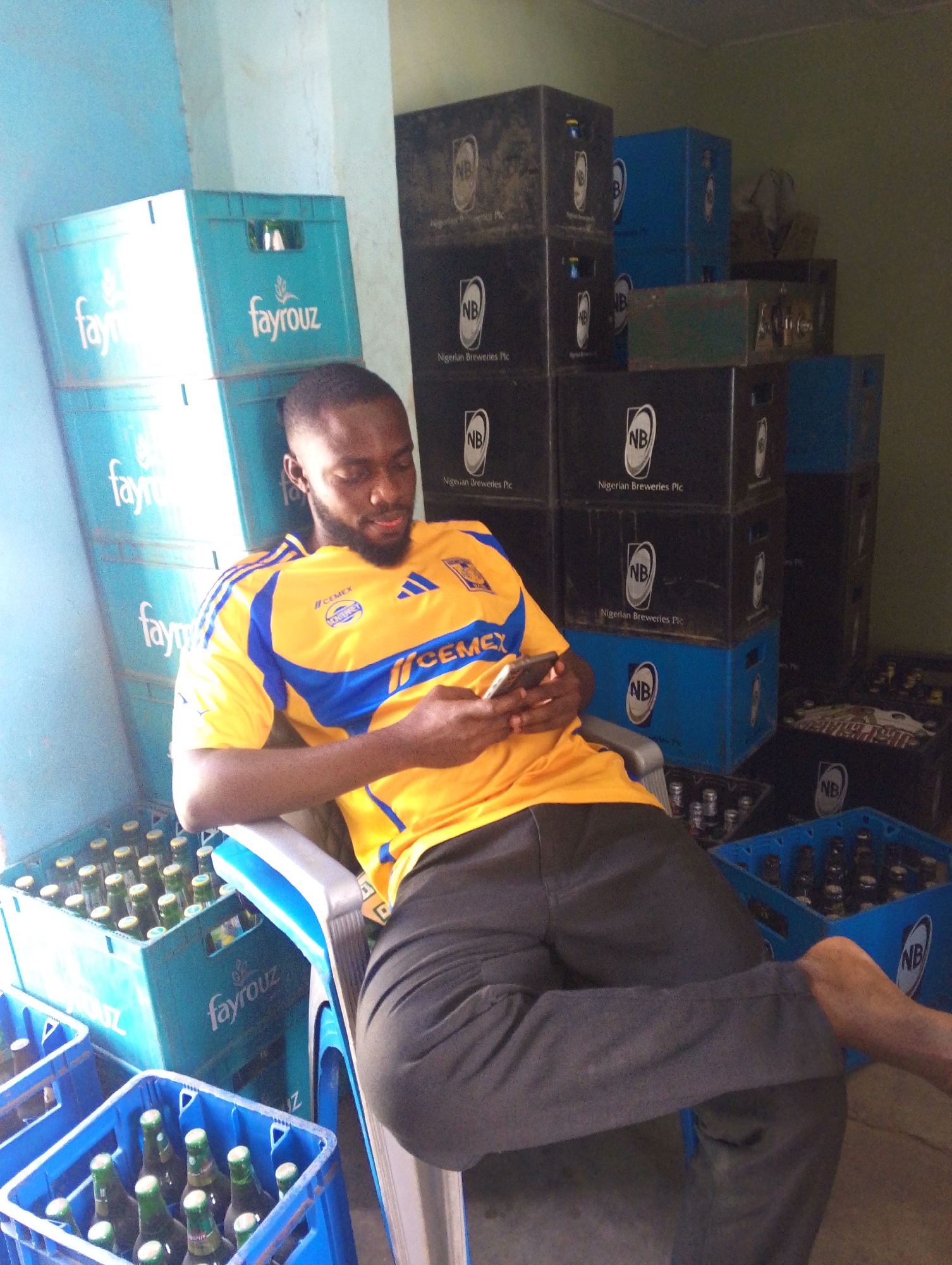 A man sits in a white plastic chair, looking at a cell phone in his hands. He wears a yellow and blue soccer jersey with the word "CEMEX" across the chest, and dark gray pants. The man is surrounded by stacks of crates, some turquoise with the word "fayrouz" printed on them, and others black and blue with the letters "NB" for Nigerian Breweries. Some of the crates are filled with green bottles. The location appears to be a storage area, likely related to a business. The city and area are unknown.